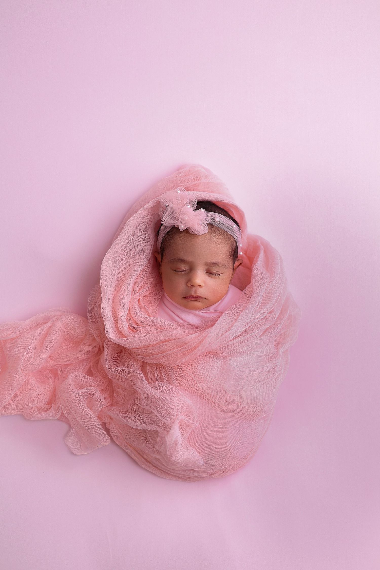 A sleeping newborn wrapped in soft pink tulle fabric against a pink background in a dreamy studio portrait series.