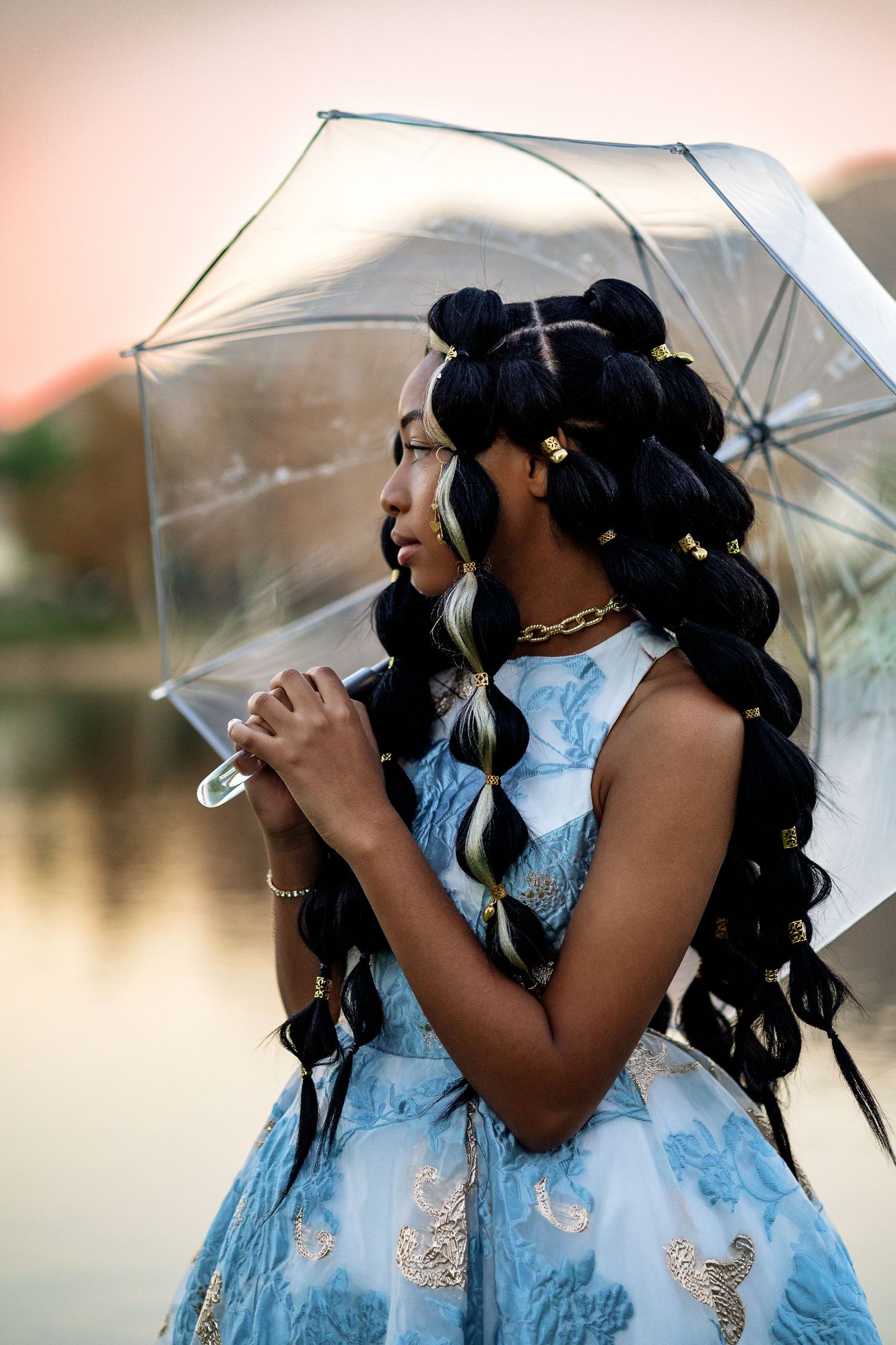 A person in a light blue formal dress poses with a clear umbrella against a soft blurred background.