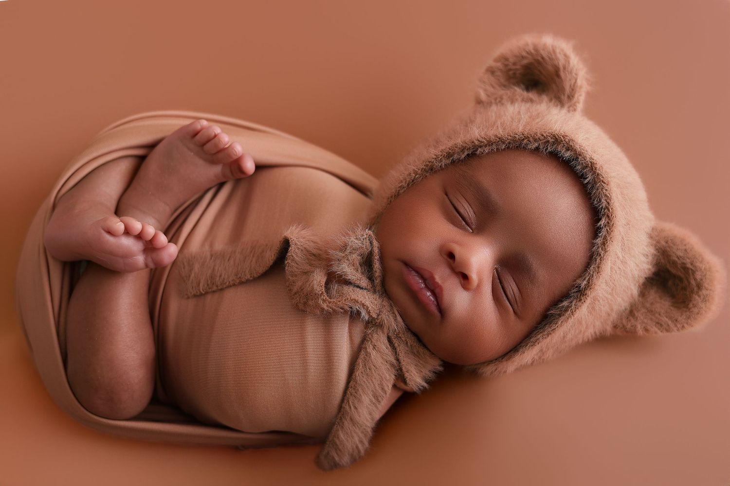 Sleeping newborn baby wearing a soft brown knit outfit curled up in peaceful poses on warm brown backdrop.