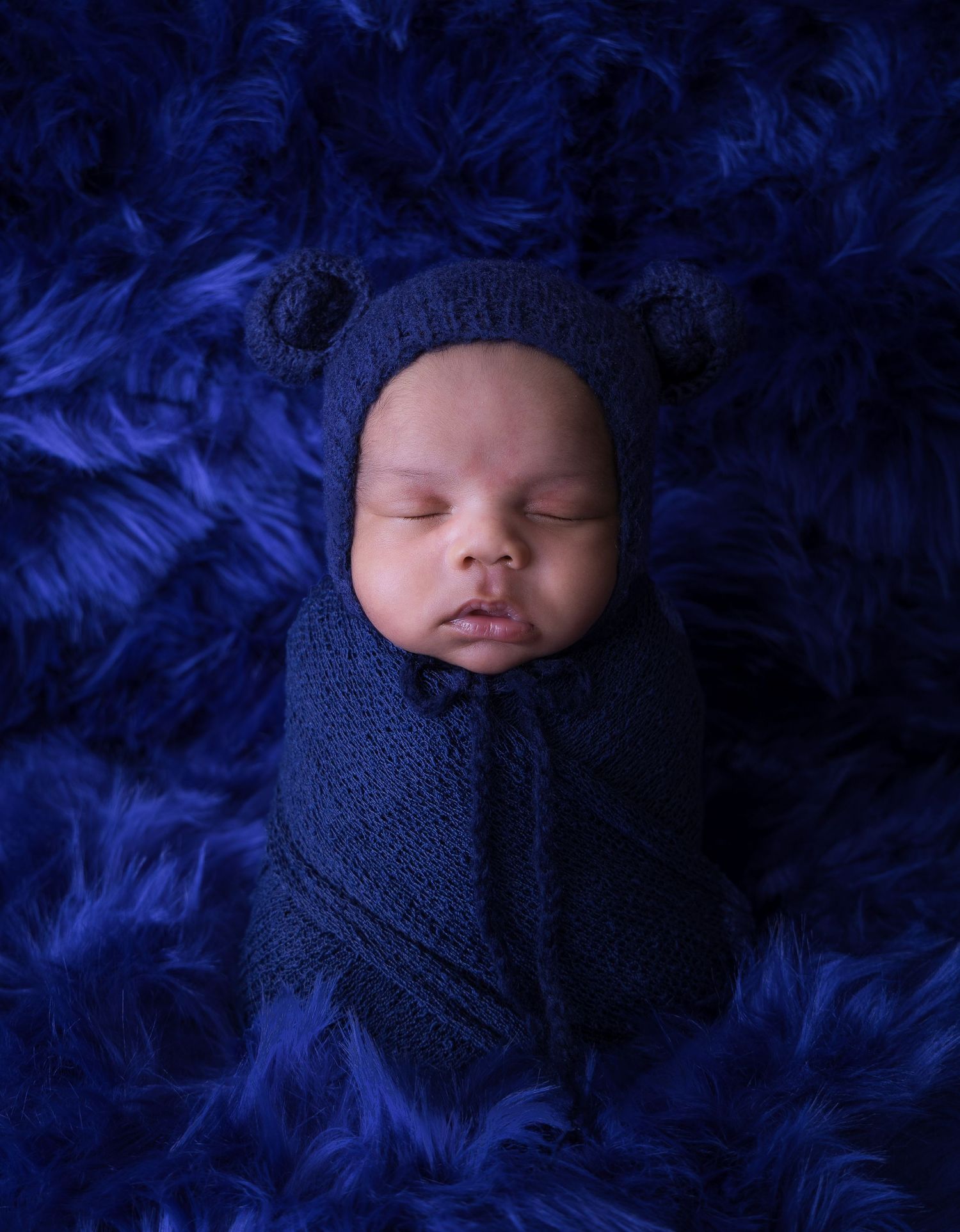 Sleeping newborn swaddled in navy blue fabric against dark blue feathered background in artistic portrait series.