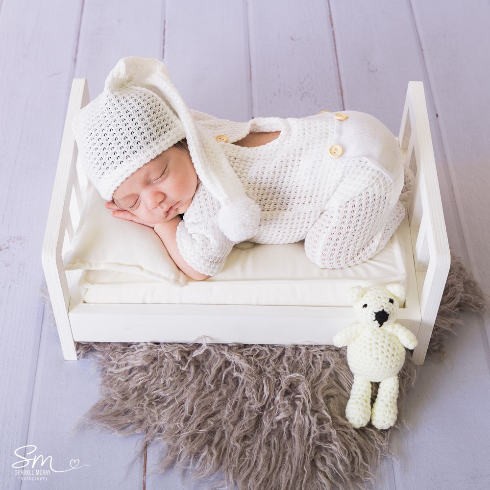 Newborn baby sleeping peacefully in white knit outfit on furry gray blanket with matching teddy bear.