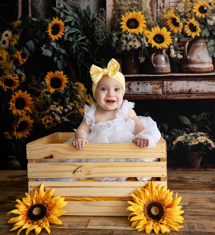 Rustic sunflower photography setup with wooden crate and yellow bow against dark floral backdrop.