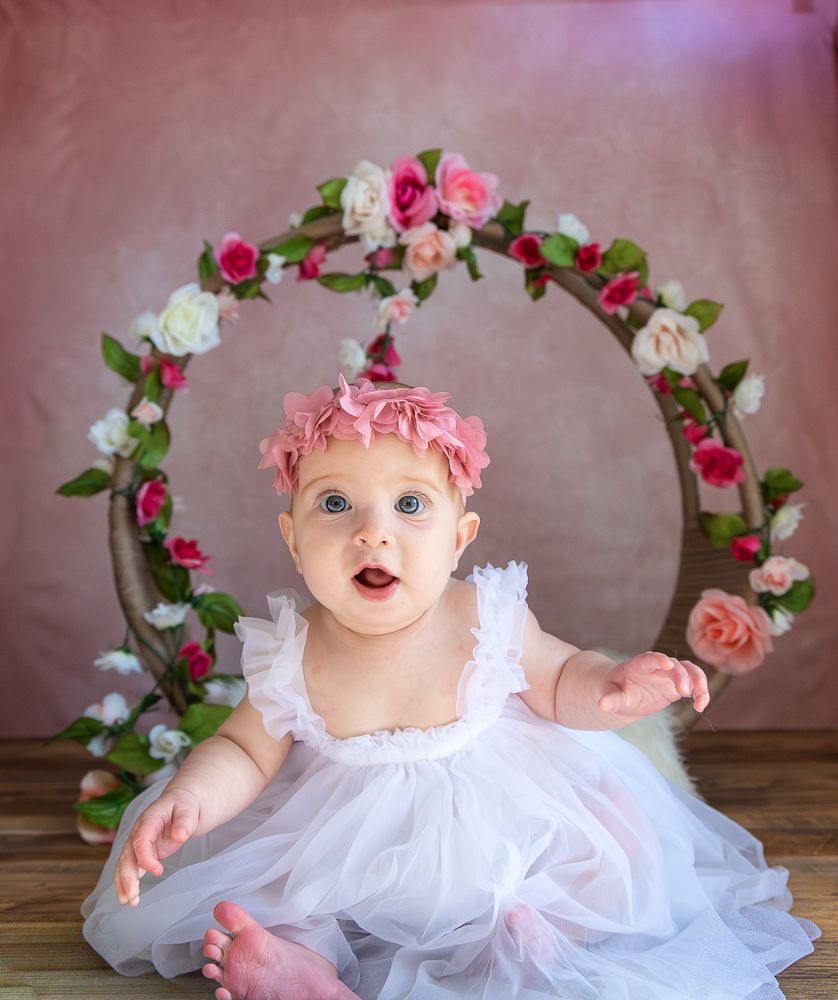 A small baby wearing a white tulle dress and pink floral crown poses on a wooden surface with floral decorations.