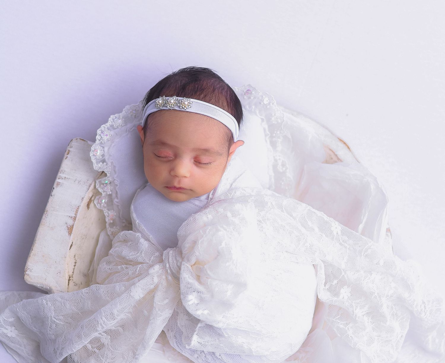 A sleeping baby wearing a white christening gown and headband lies peacefully against a light background.