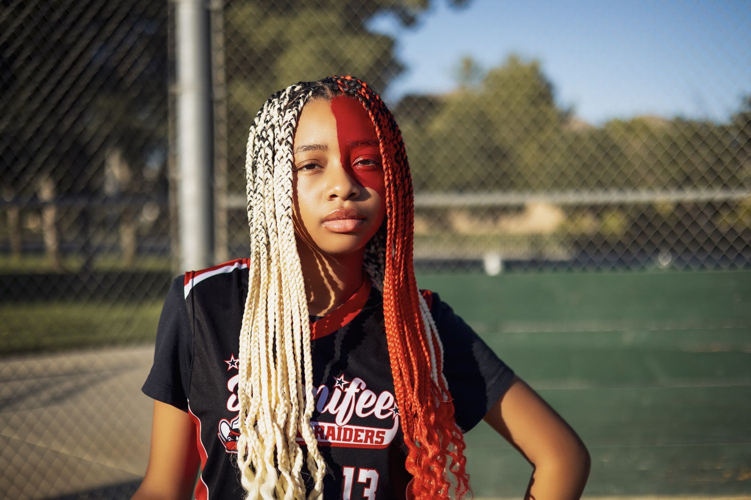 Softball player with blonde and red braided hair wearing black team jersey stands on field during golden hour.