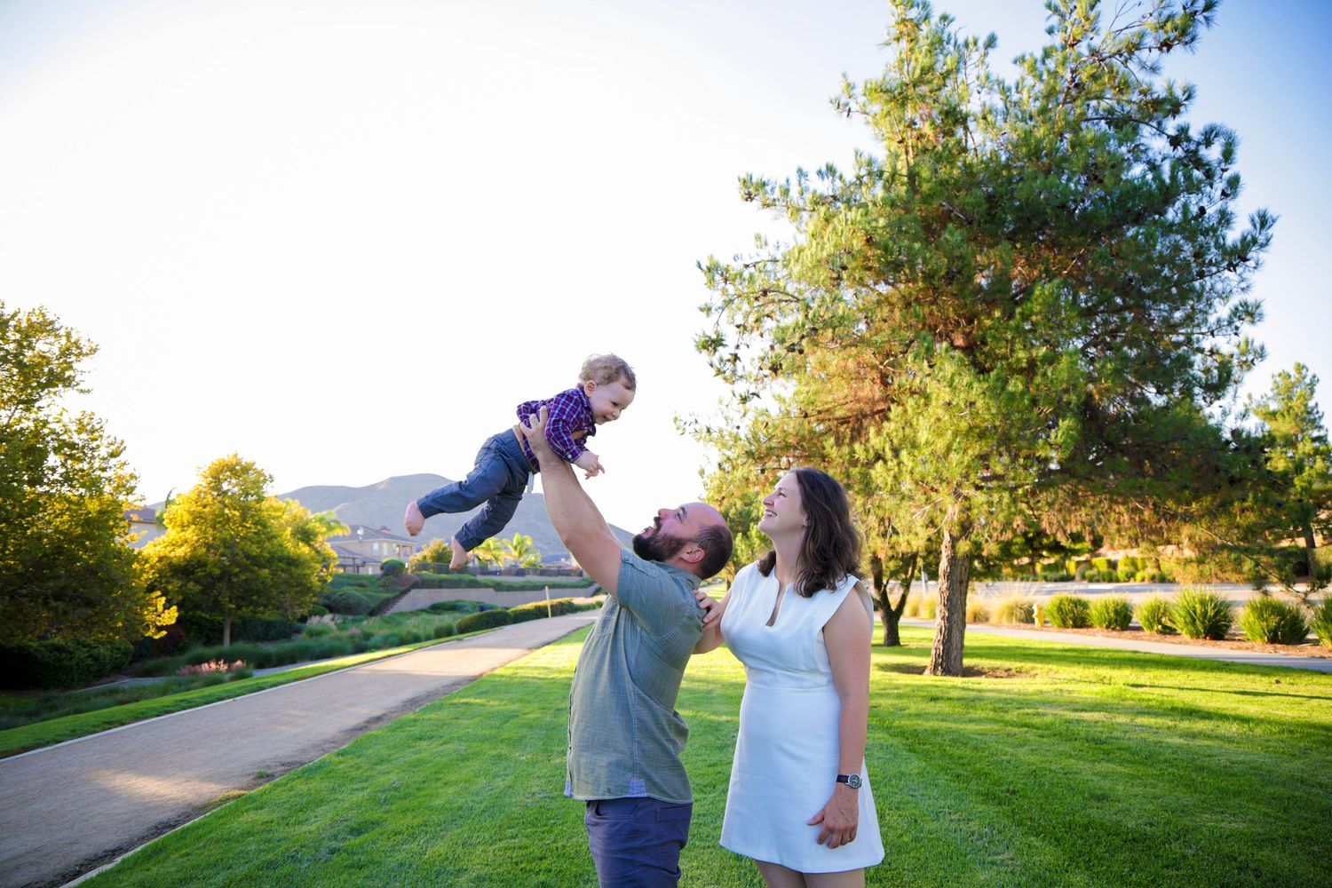 A couple plays with their child in a sunny park with mountains in the background during golden hour.