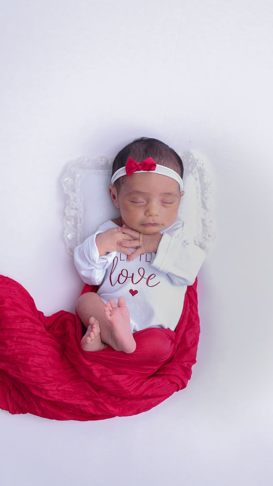 Newborn baby sleeping peacefully wrapped in red fabric with white outfit and small red bow headband.