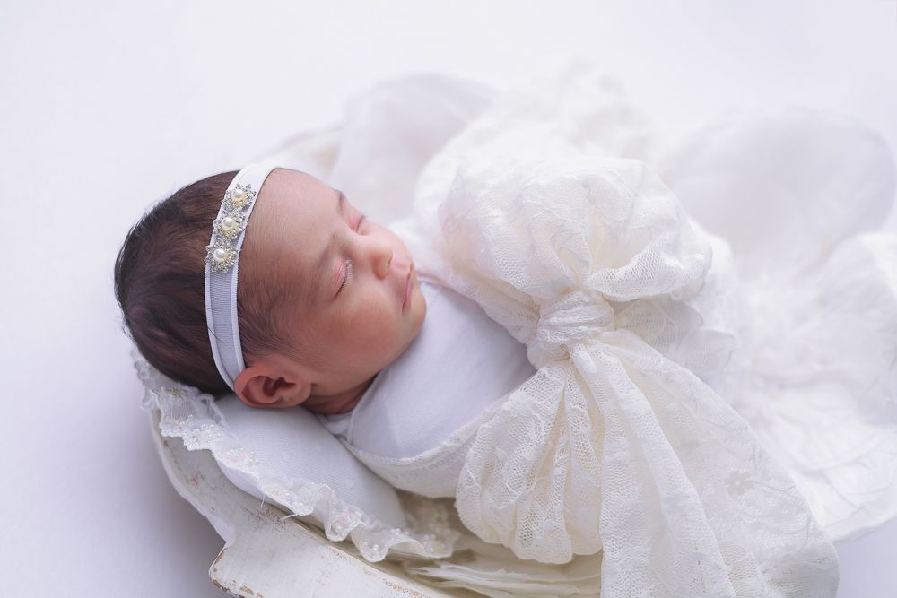 A sleeping newborn baby in white lace dress and headband lies peacefully on white bedding.