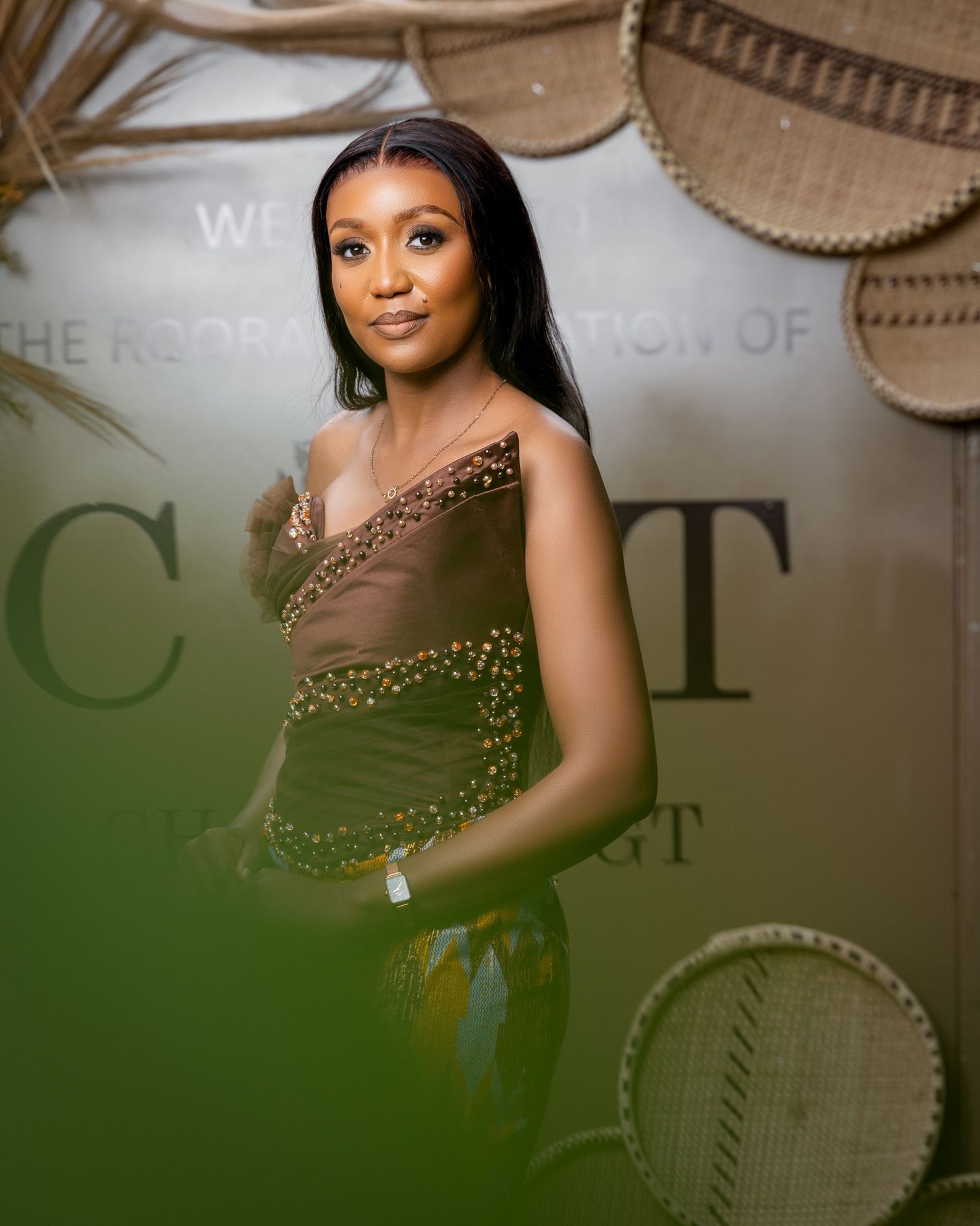 Woman in elegant olive green embellished outfit poses at an upscale event with woven basket decor backdrop.