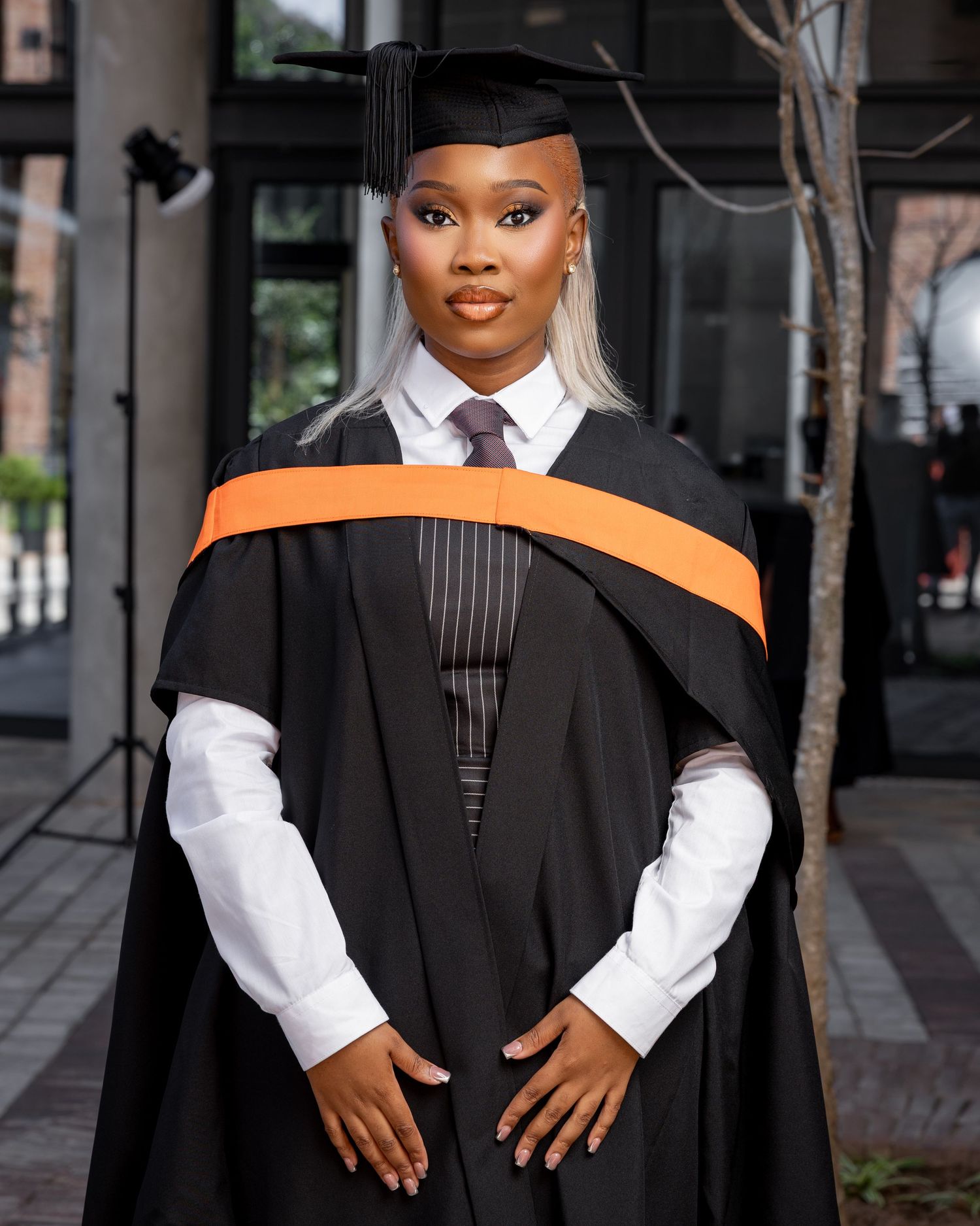 Graduate in black cap and gown with orange sash poses proudly at outdoor graduation ceremony.