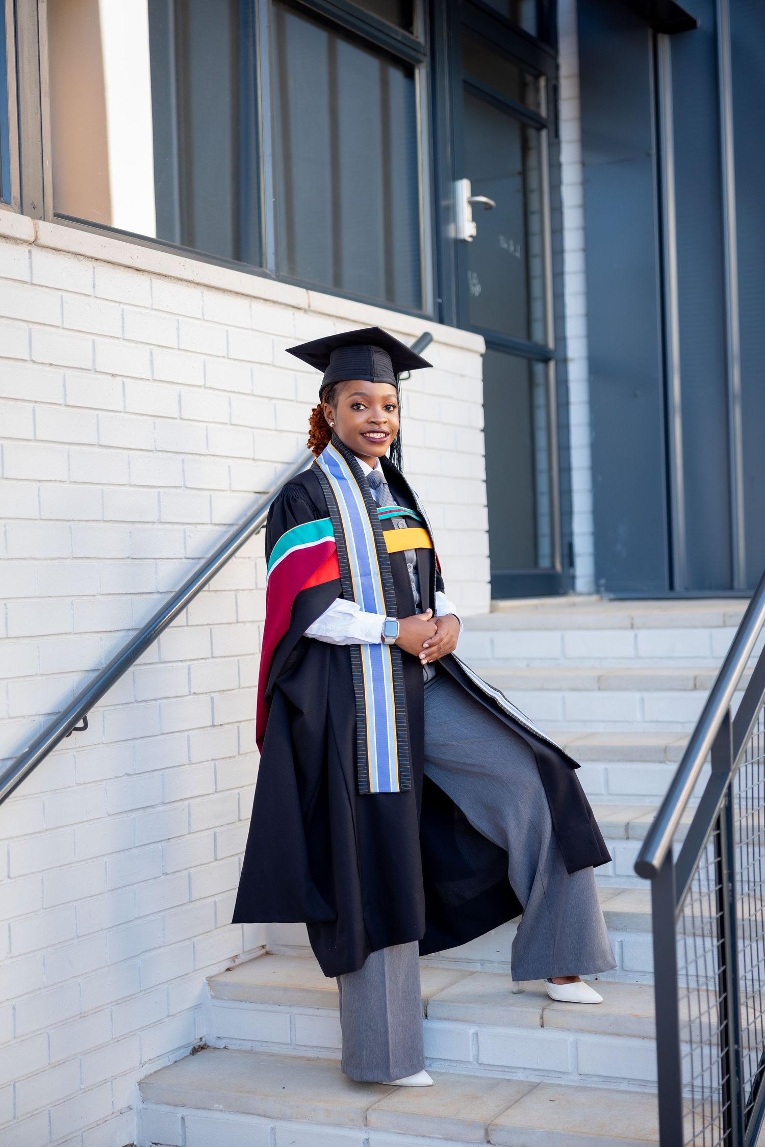 A young Black woman in graduation gown, cap, and colorful stole stands proudly on outdoor steps holding her diploma.