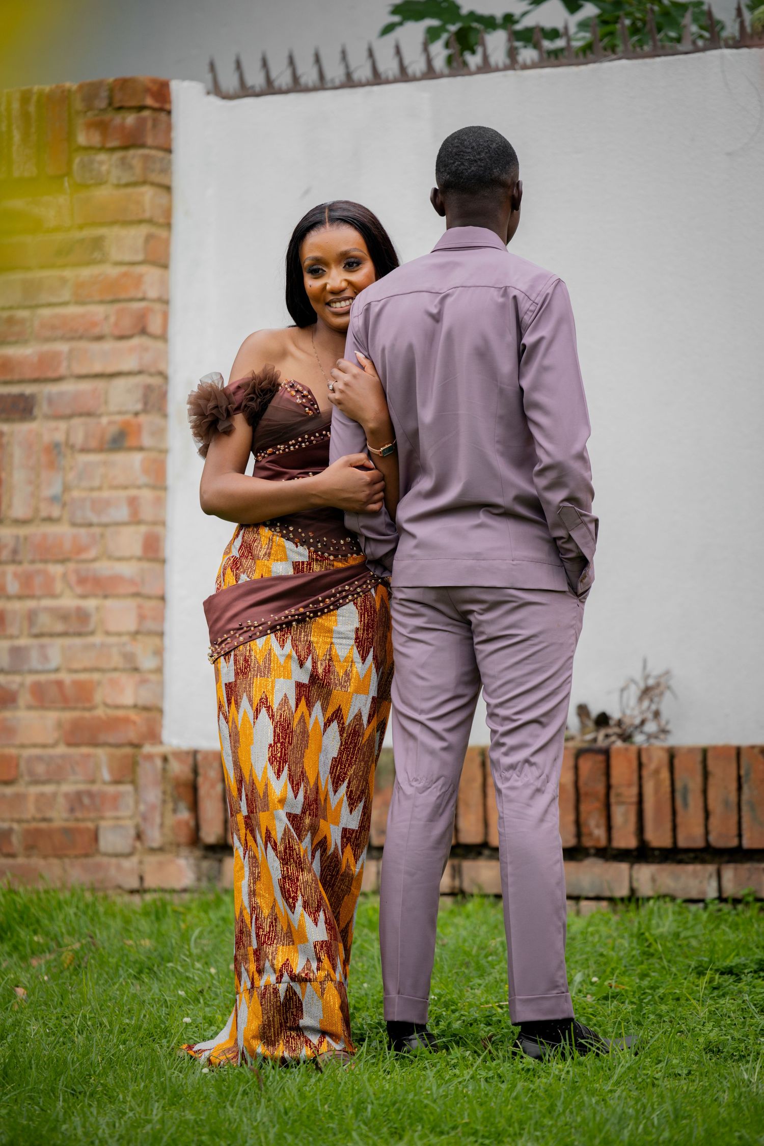Couple in traditional African attire embracing outdoors, woman in colorful kente dress smiling at camera, man in lavender suit.