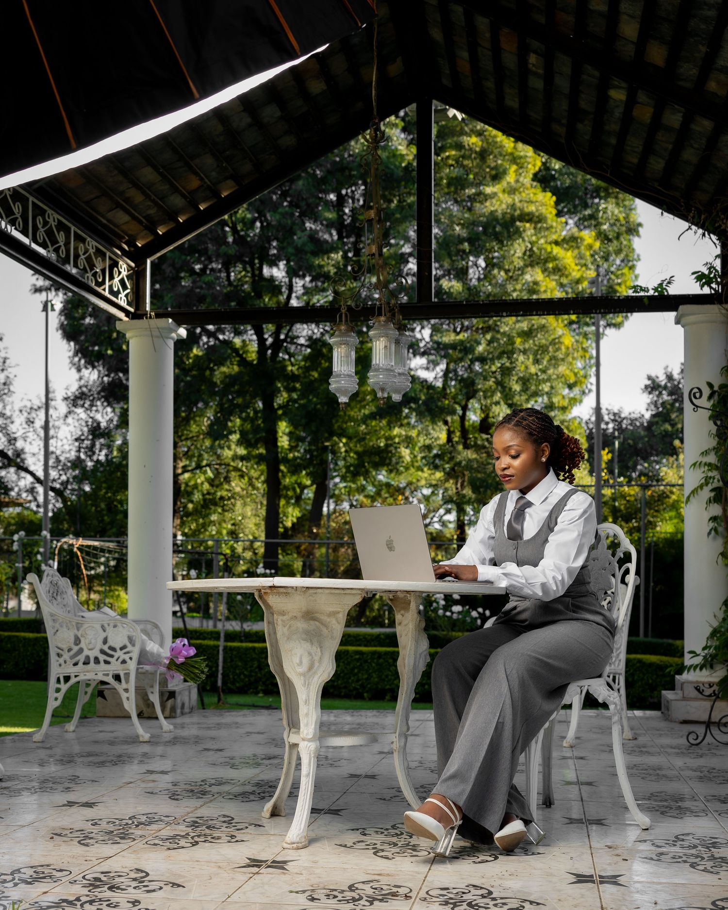 A professional woman in gray vest and trousers works on a laptop at an ornate white table under an elegant outdoor gazebo.