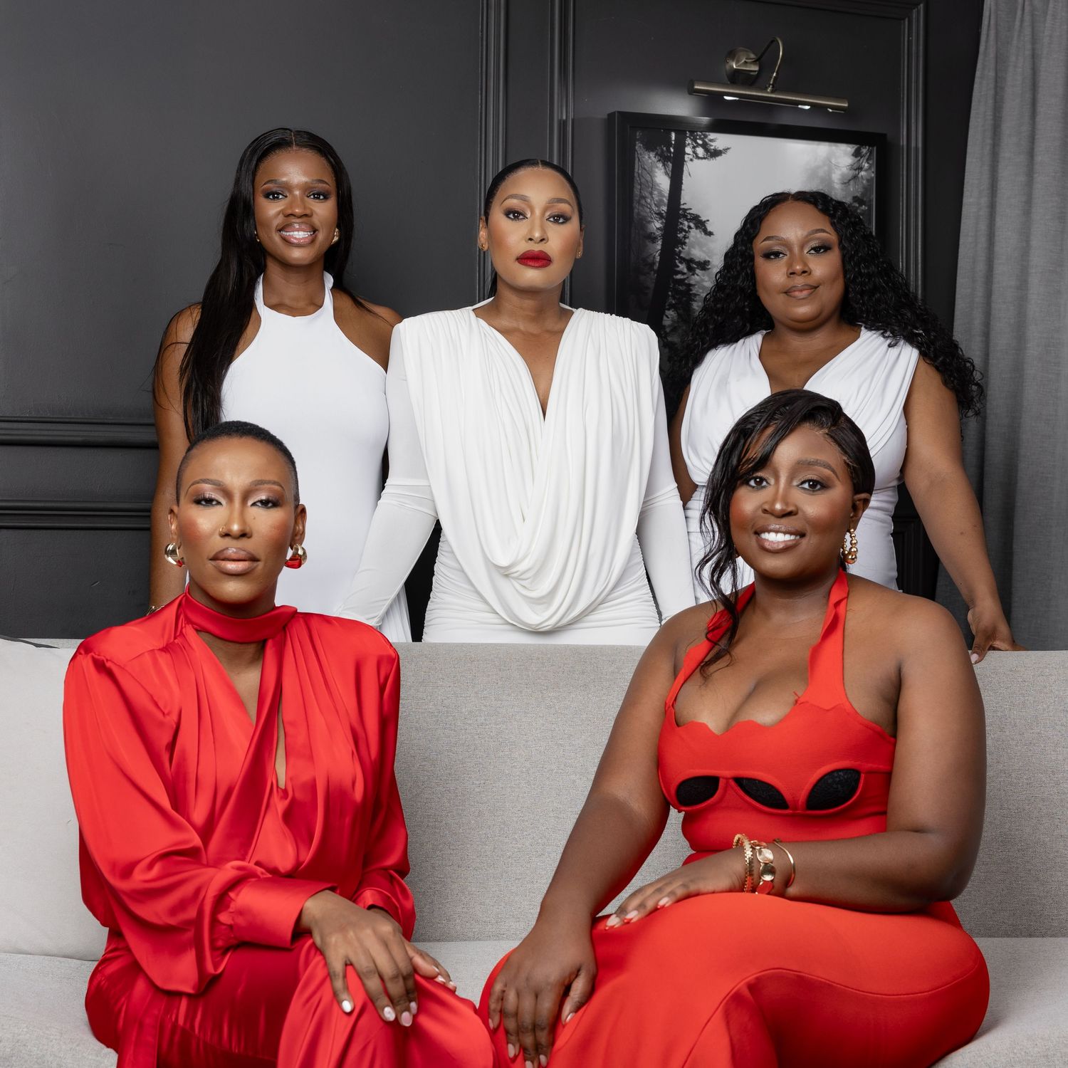 Five elegant women posing together, three in white outfits and two in red, in a stylish modern interior setting.