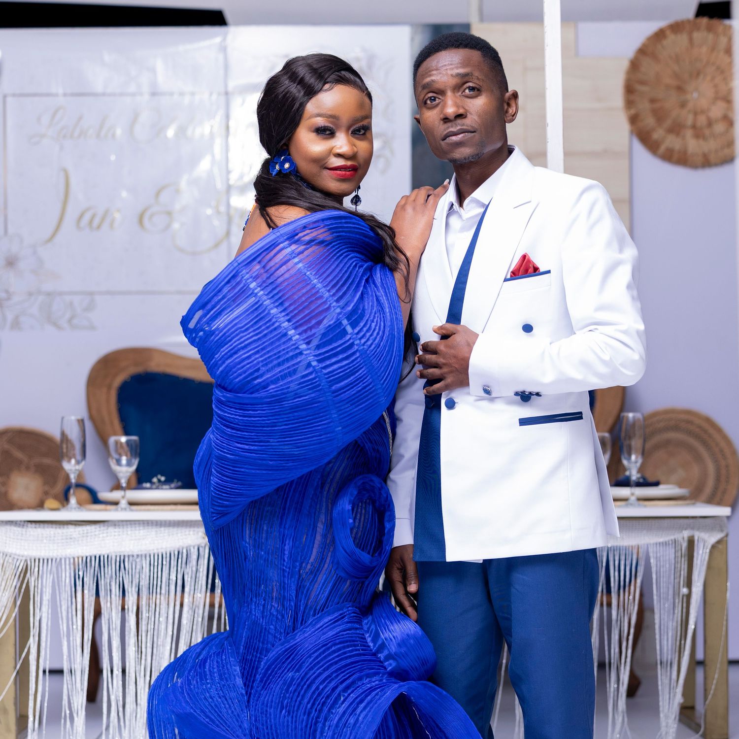 Couple in royal blue gown and white suit pose together at decorated event venue.