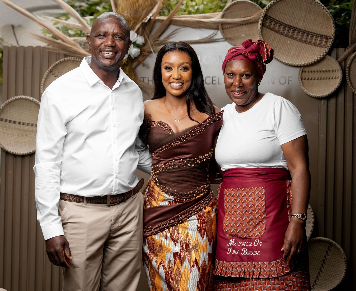 Three people in traditional African attire smile together at a celebratory event with woven baskets on the wall.