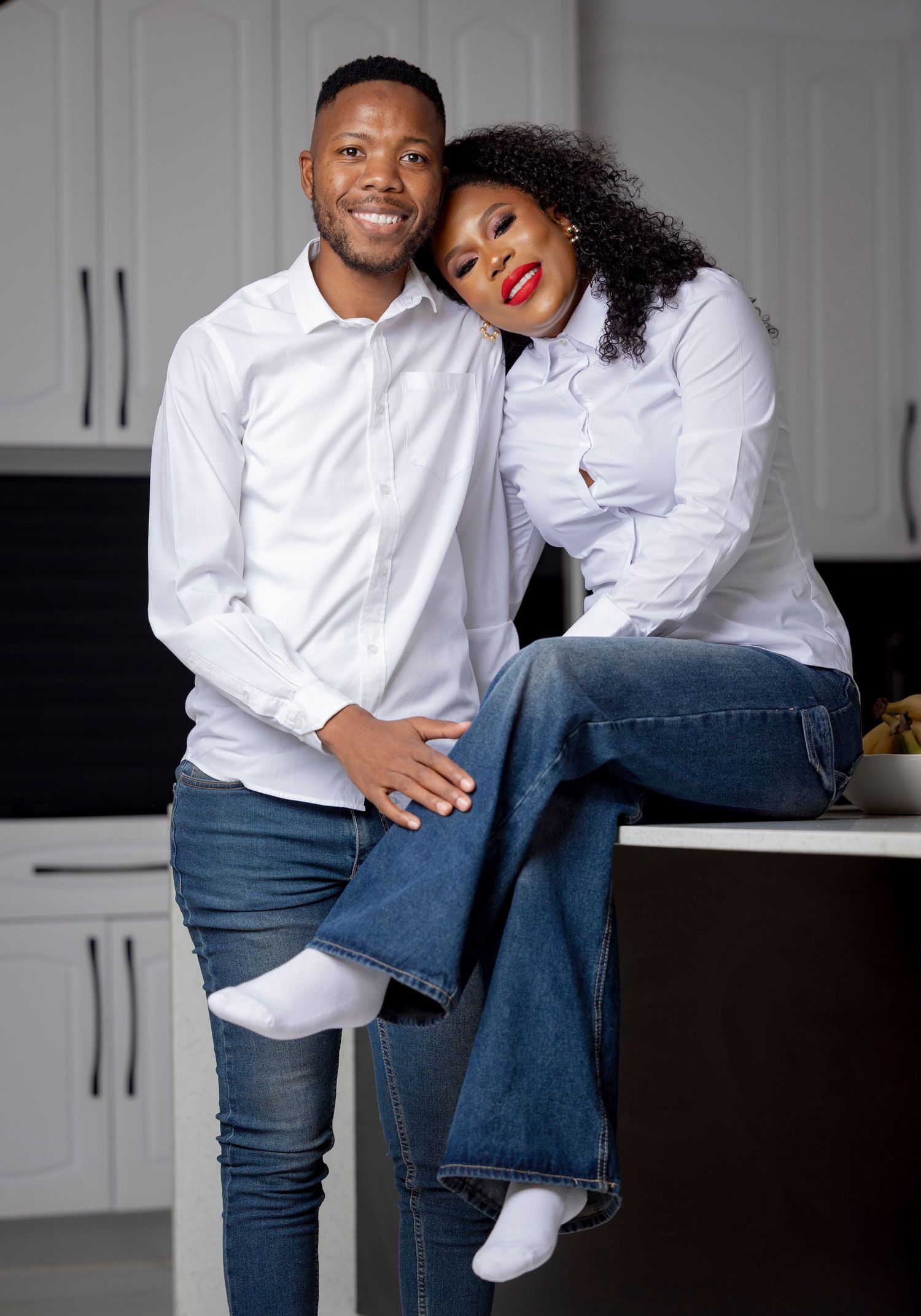 A couple in matching white shirts and blue jeans pose playfully together in a modern kitchen setting.