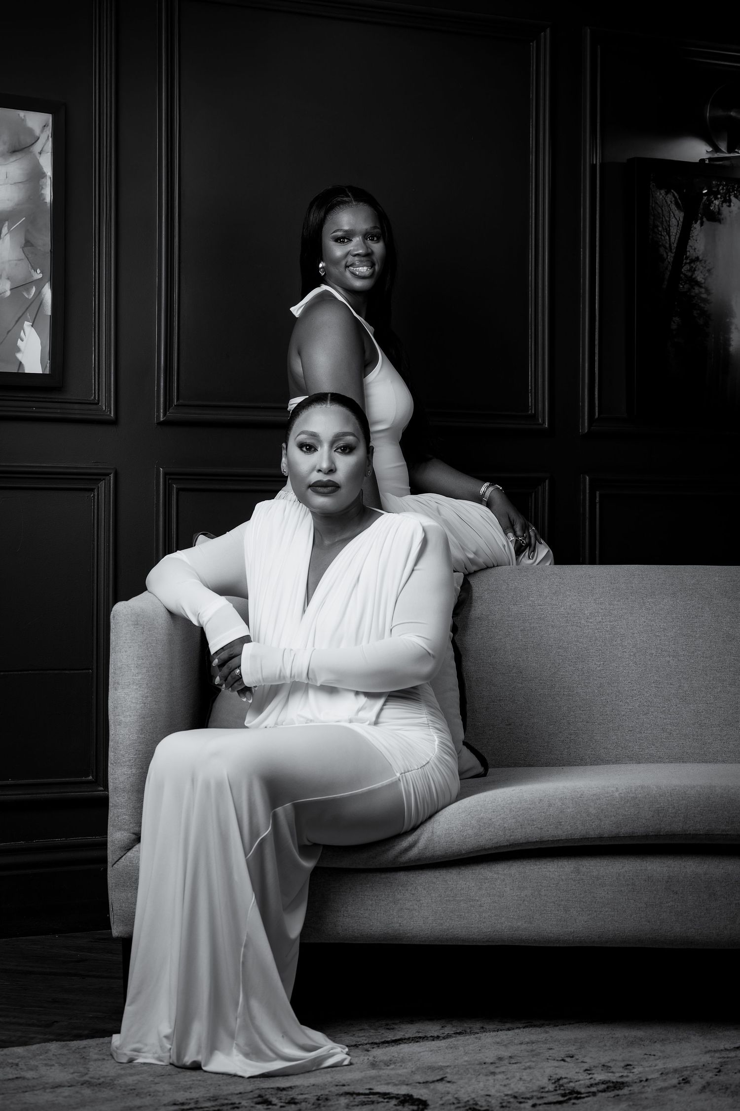 Two women in white outfits pose elegantly on a gray sofa in a black and white portrait photo.