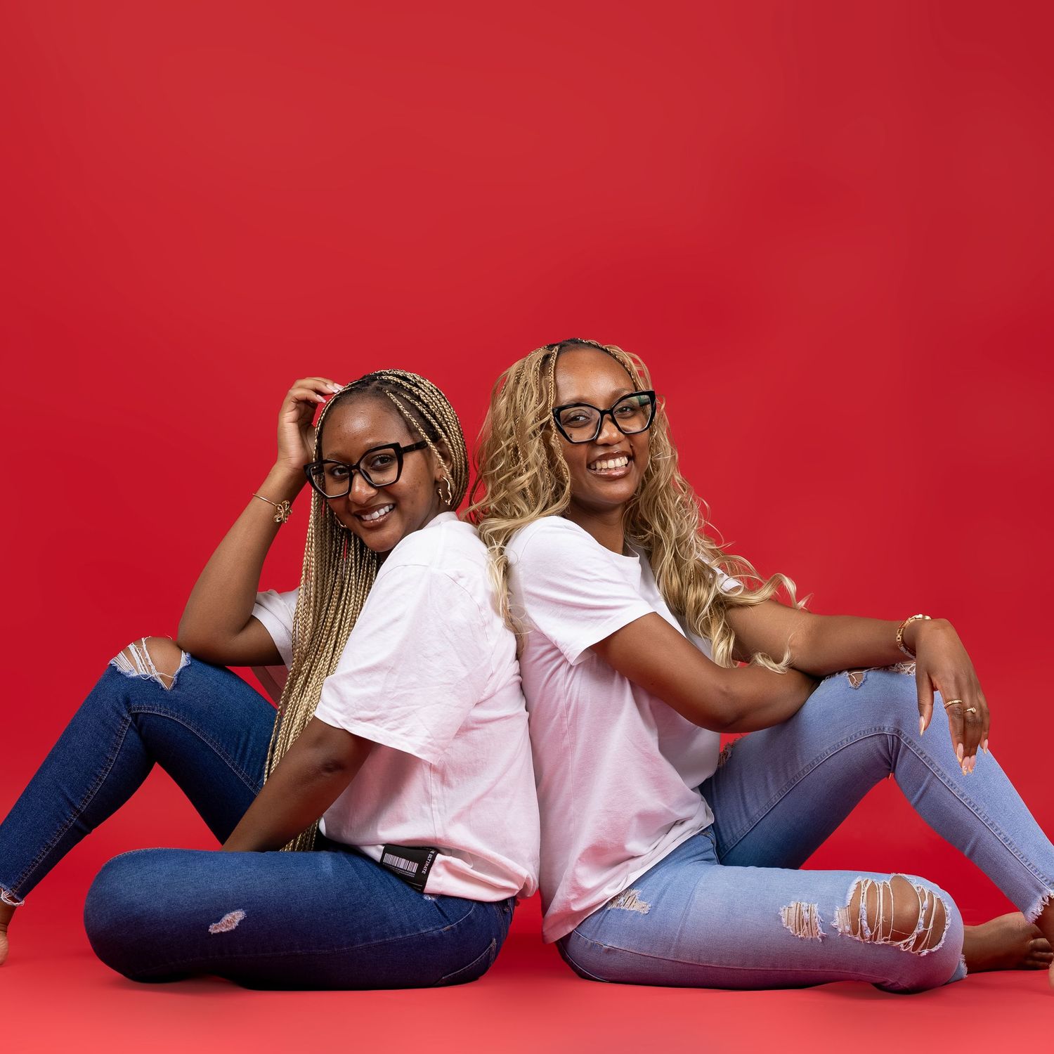 Two young women wearing glasses and jeans sit back-to-back smiling against a vibrant red background.