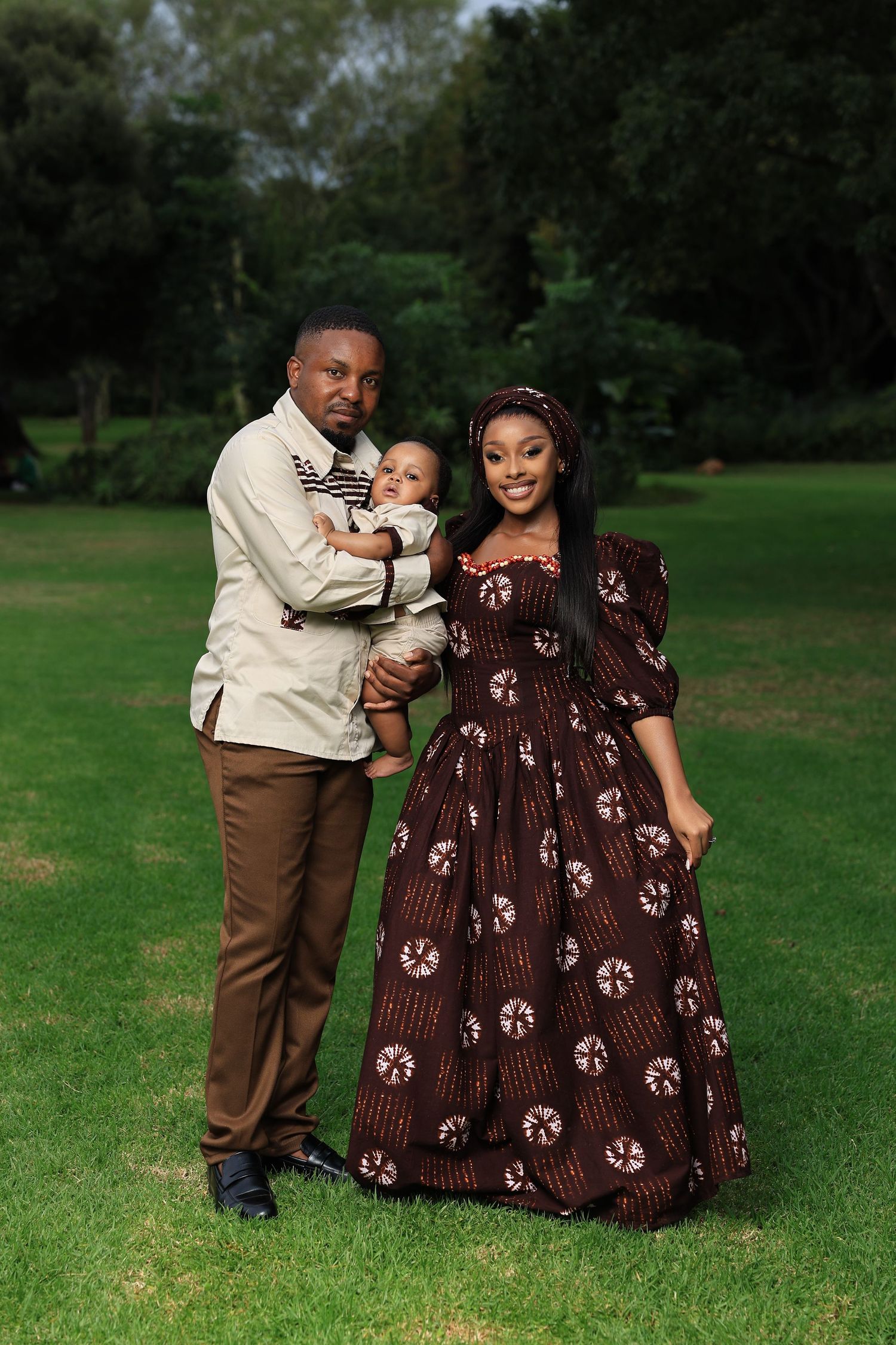 A family portrait of a couple in African attire holding their baby on a lush green lawn outdoors.