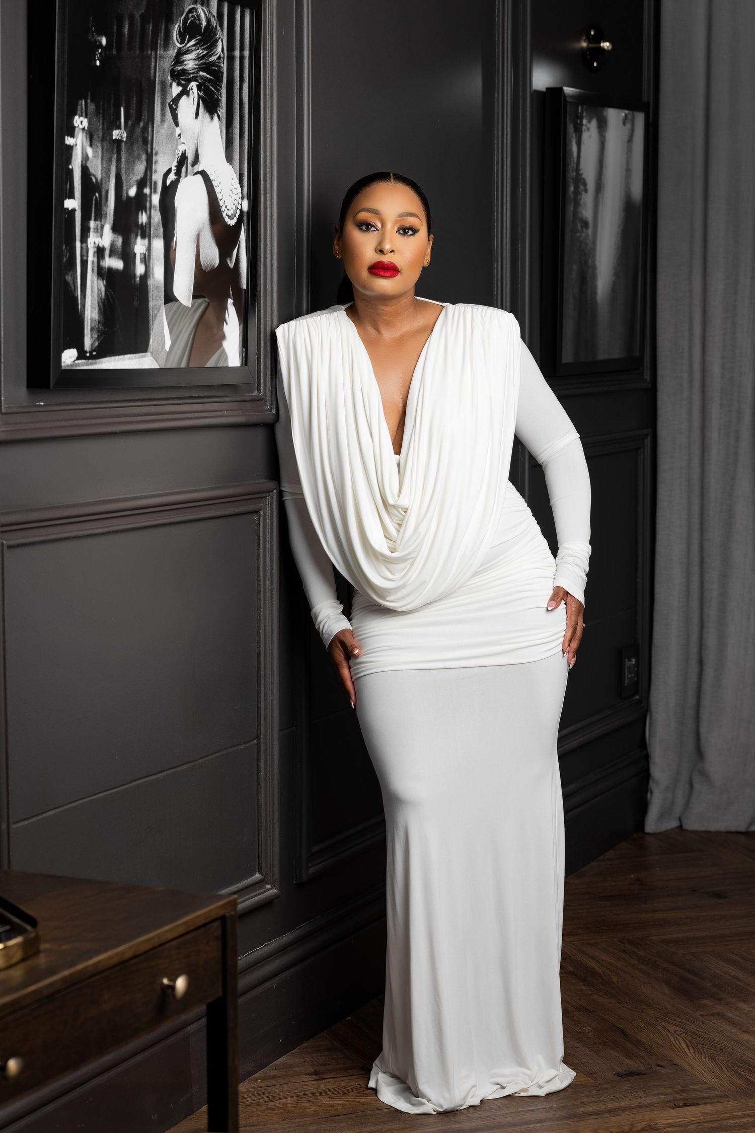 Woman in elegant white cowl-neck gown with deep V-neckline poses in sophisticated dark interior setting.