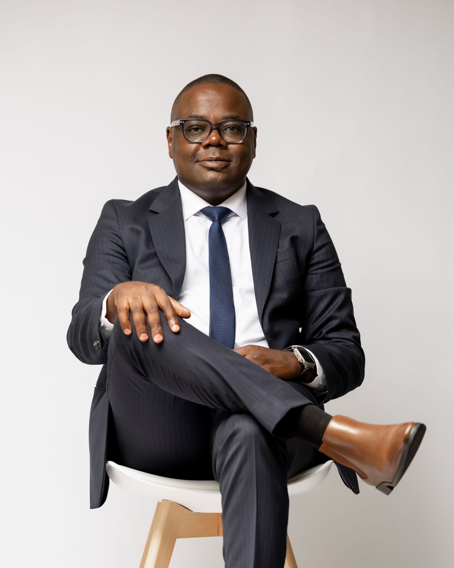 Professional in a dark suit and glasses sits confidently on a wooden chair against a light background.
