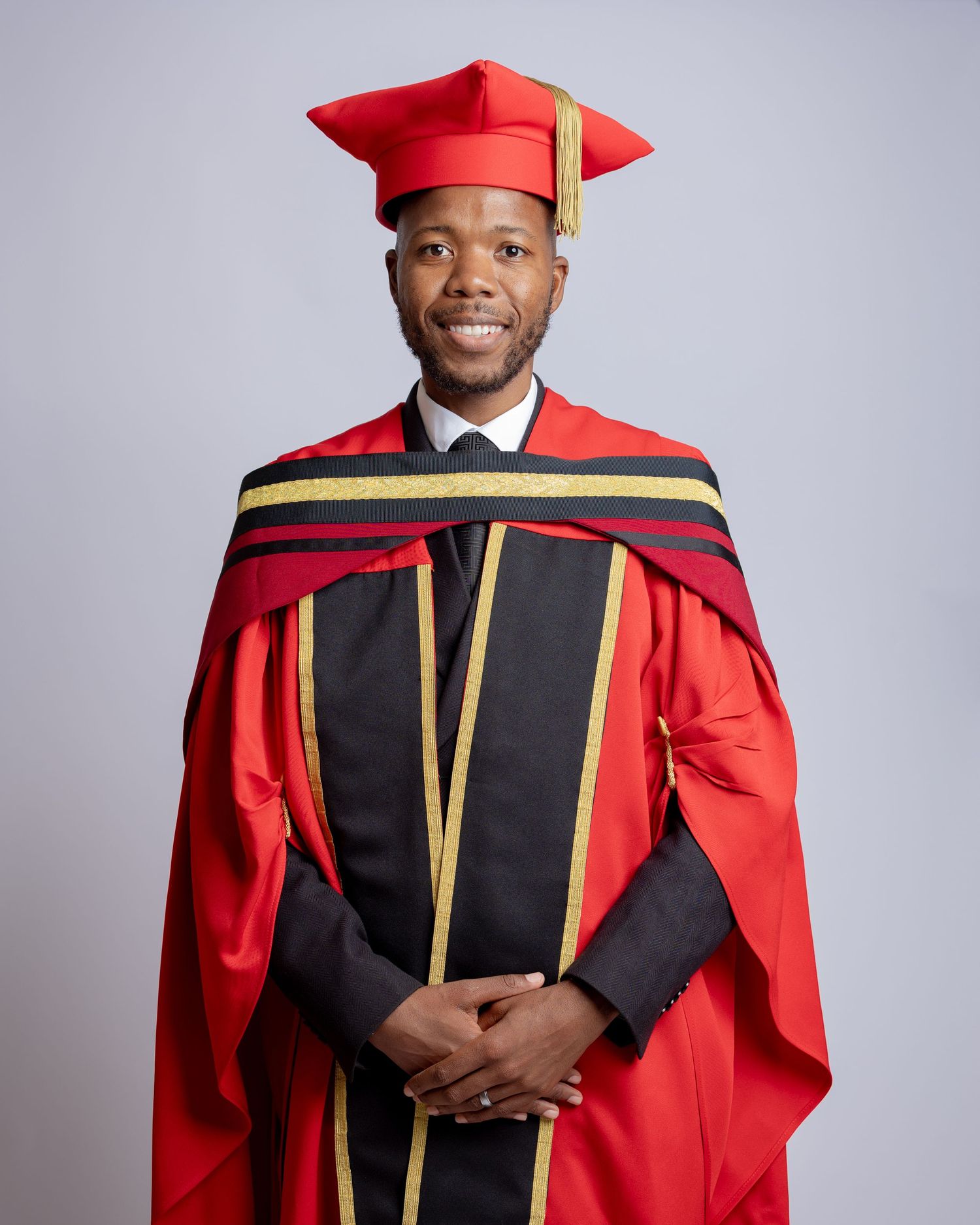 Graduate wearing red academic regalia with gold trim and black panels poses against grey background for portraits.