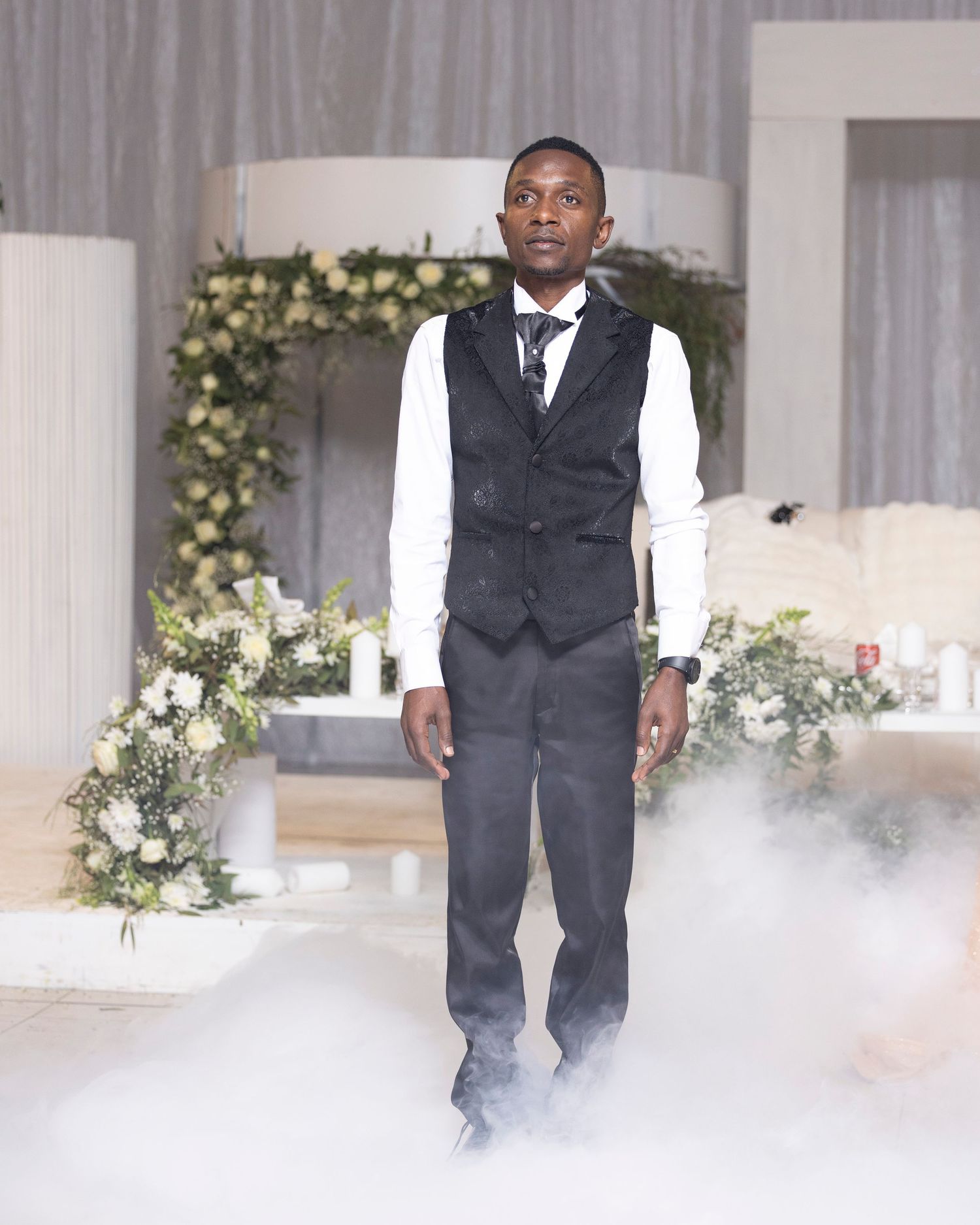 Wedding party posing in formal attire against white backdrop with floral decor.