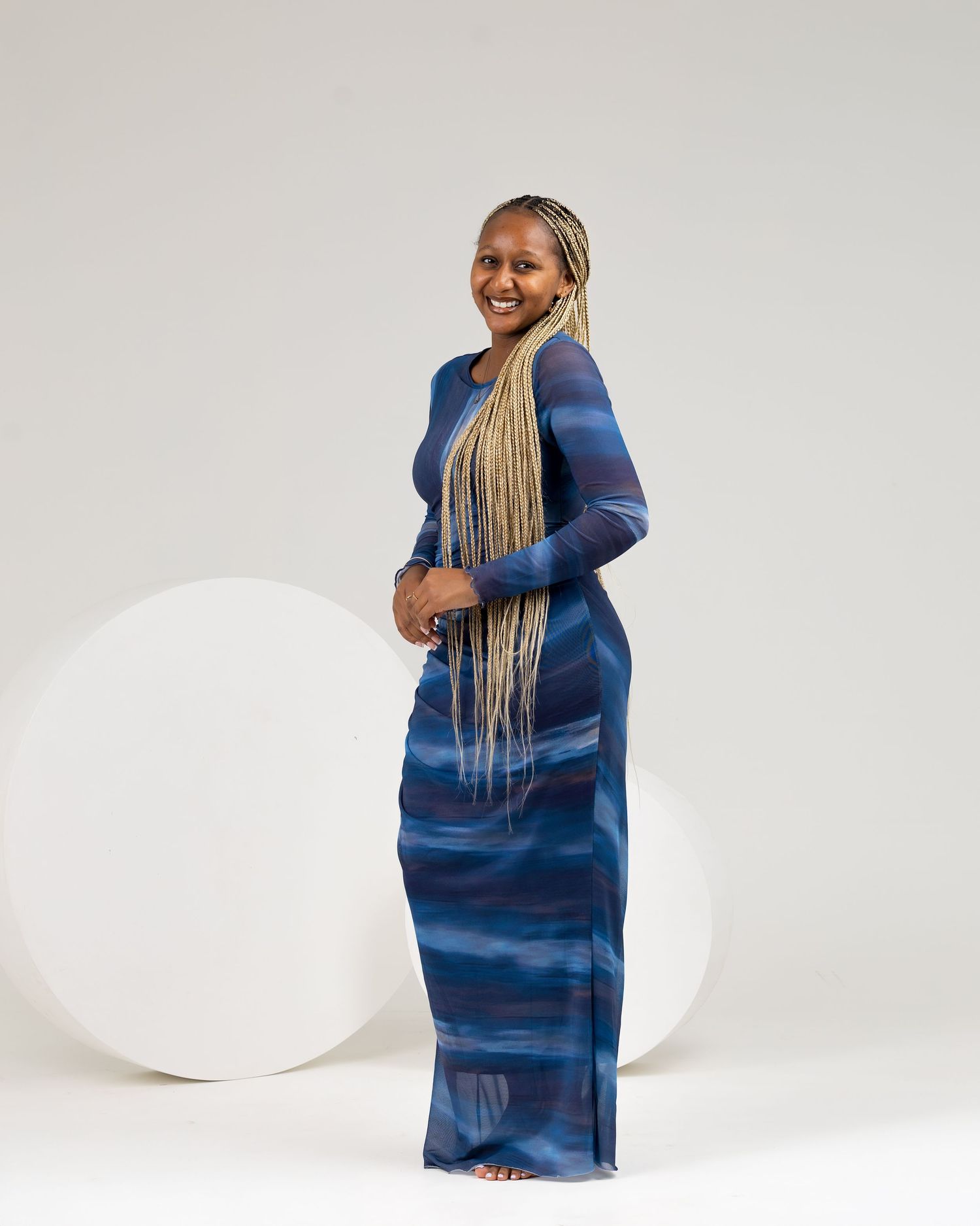 Woman in blue patterned maxi dress with long blonde braids smiling, posing sideways against white background.