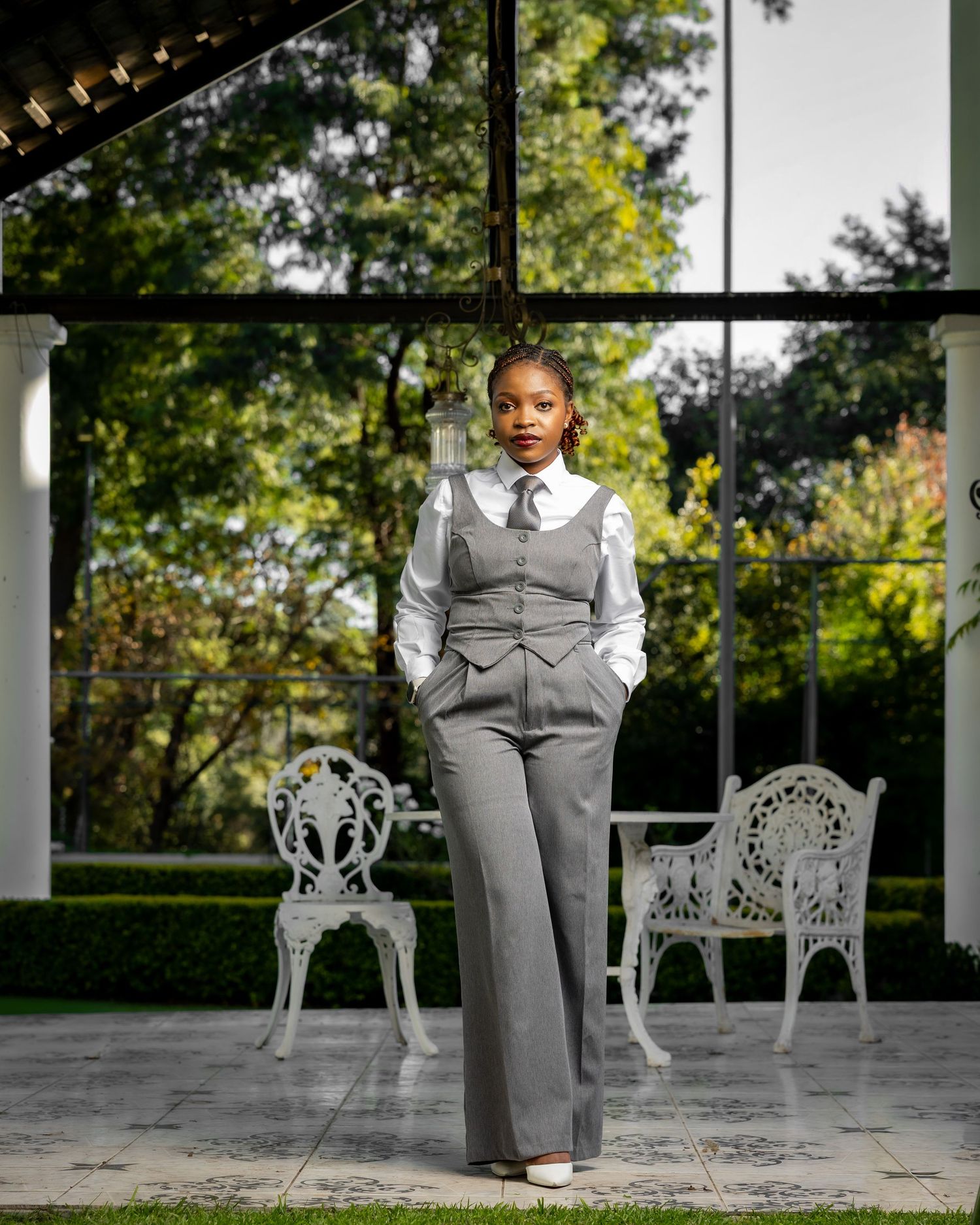 A confident woman poses in a gray vest pantsuit with white shirt and tie, standing in an elegant glass-walled venue.