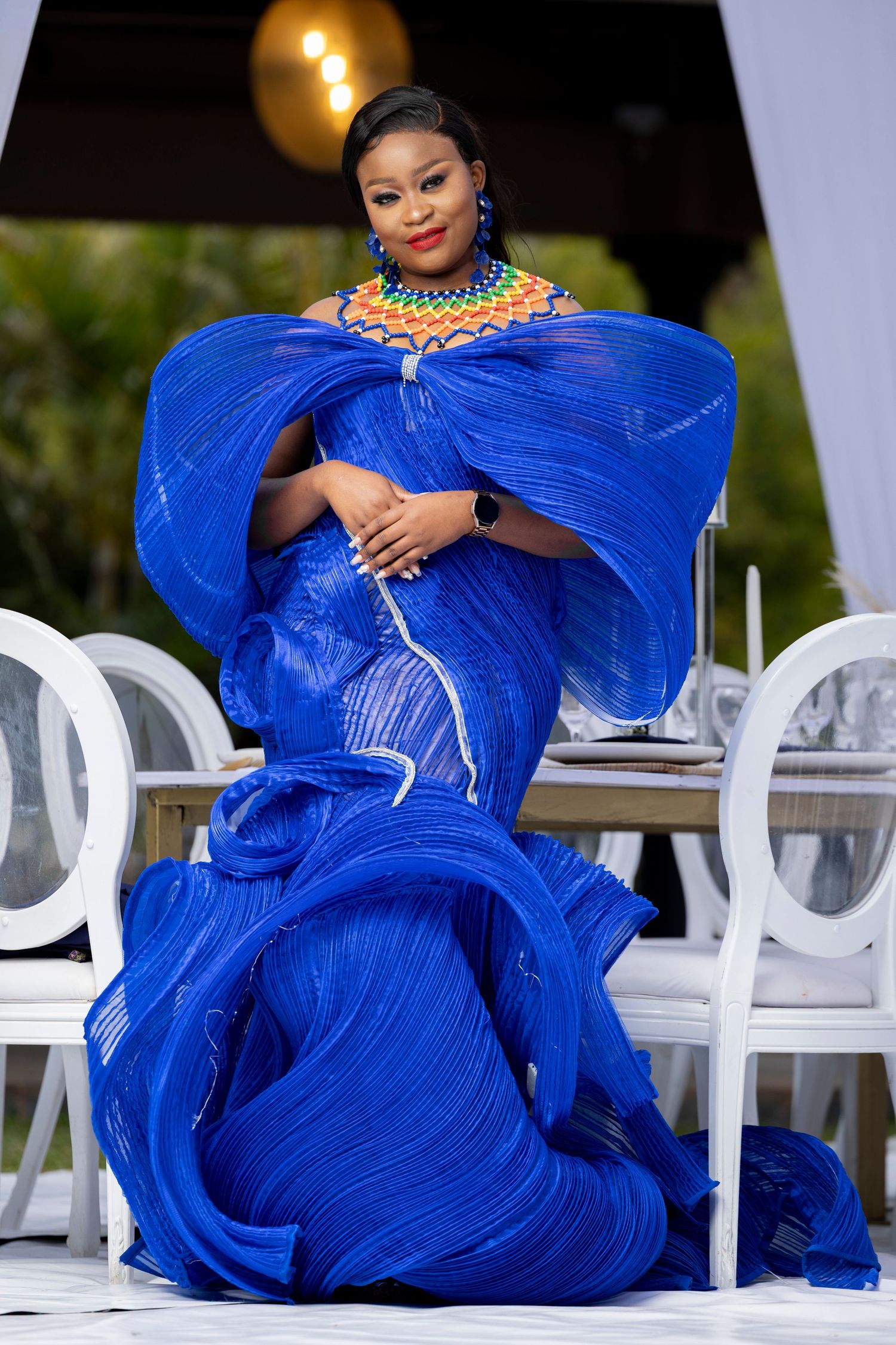 Person in flowing royal blue gown poses dramatically on white outdoor furniture.