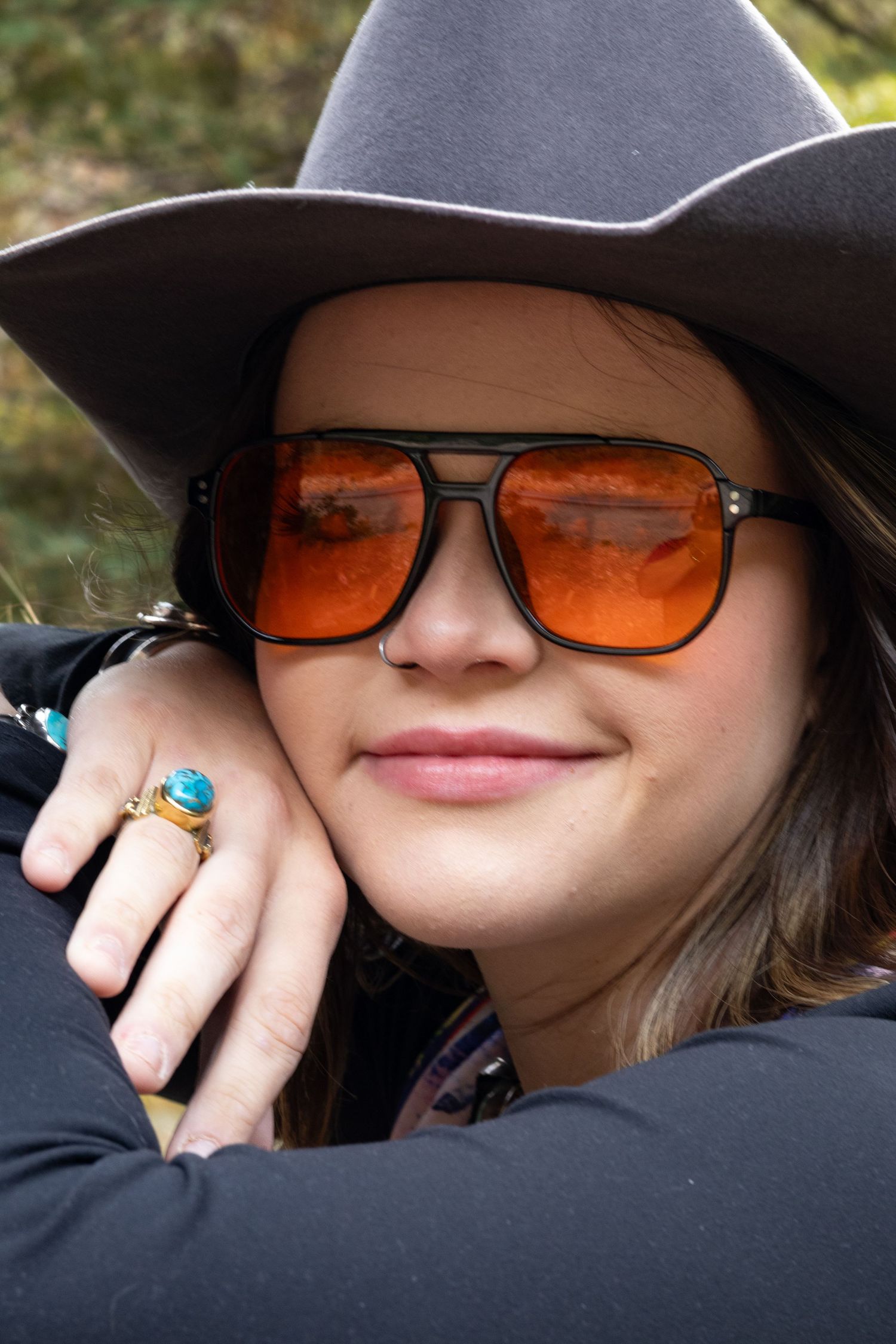 Young woman wearing orange tinted aviator sunglasses and a gray cowboy hat smiles in an outdoor setting.