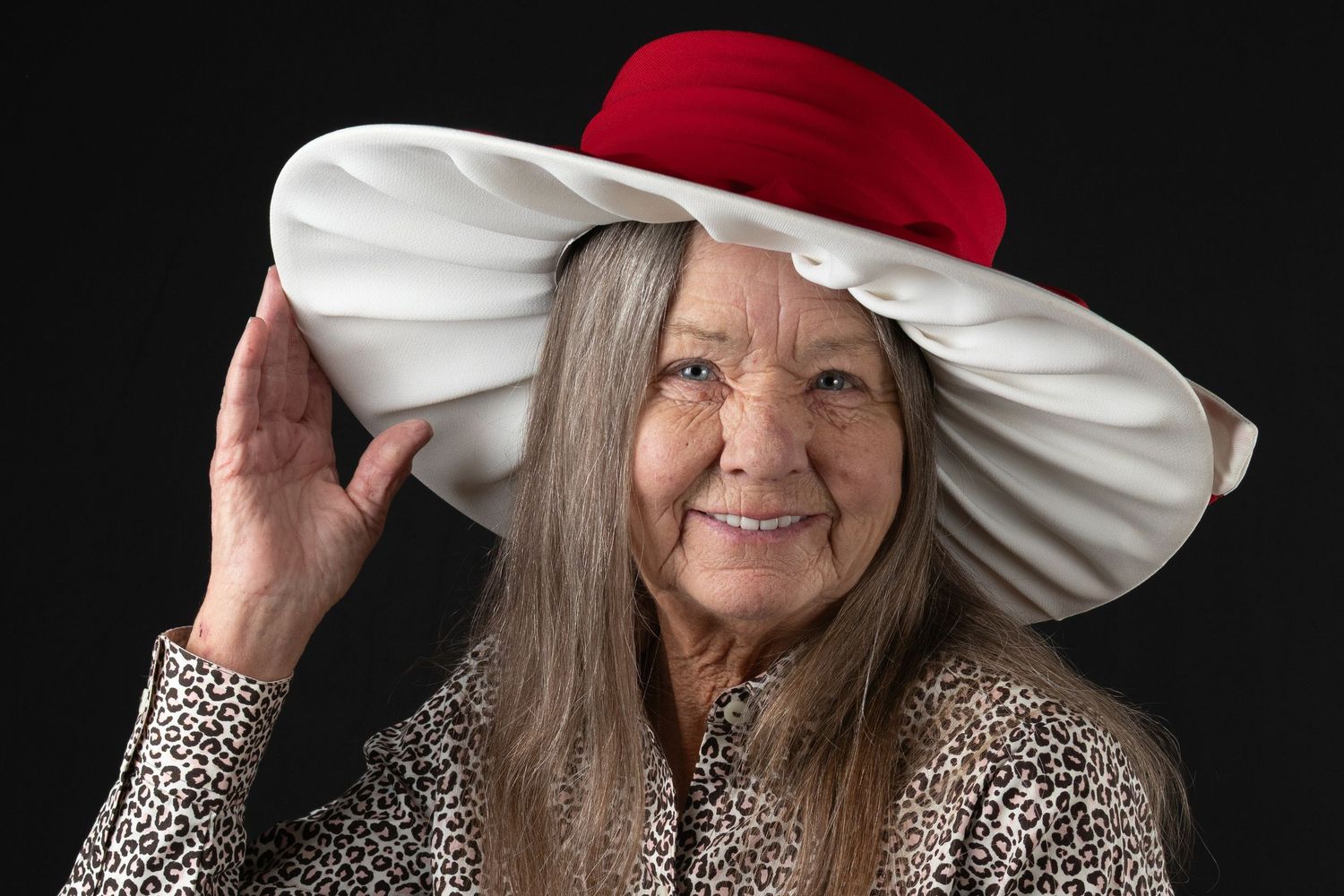 Cheerful elderly woman with long gray hair wearing large red and white decorative hat on black background.