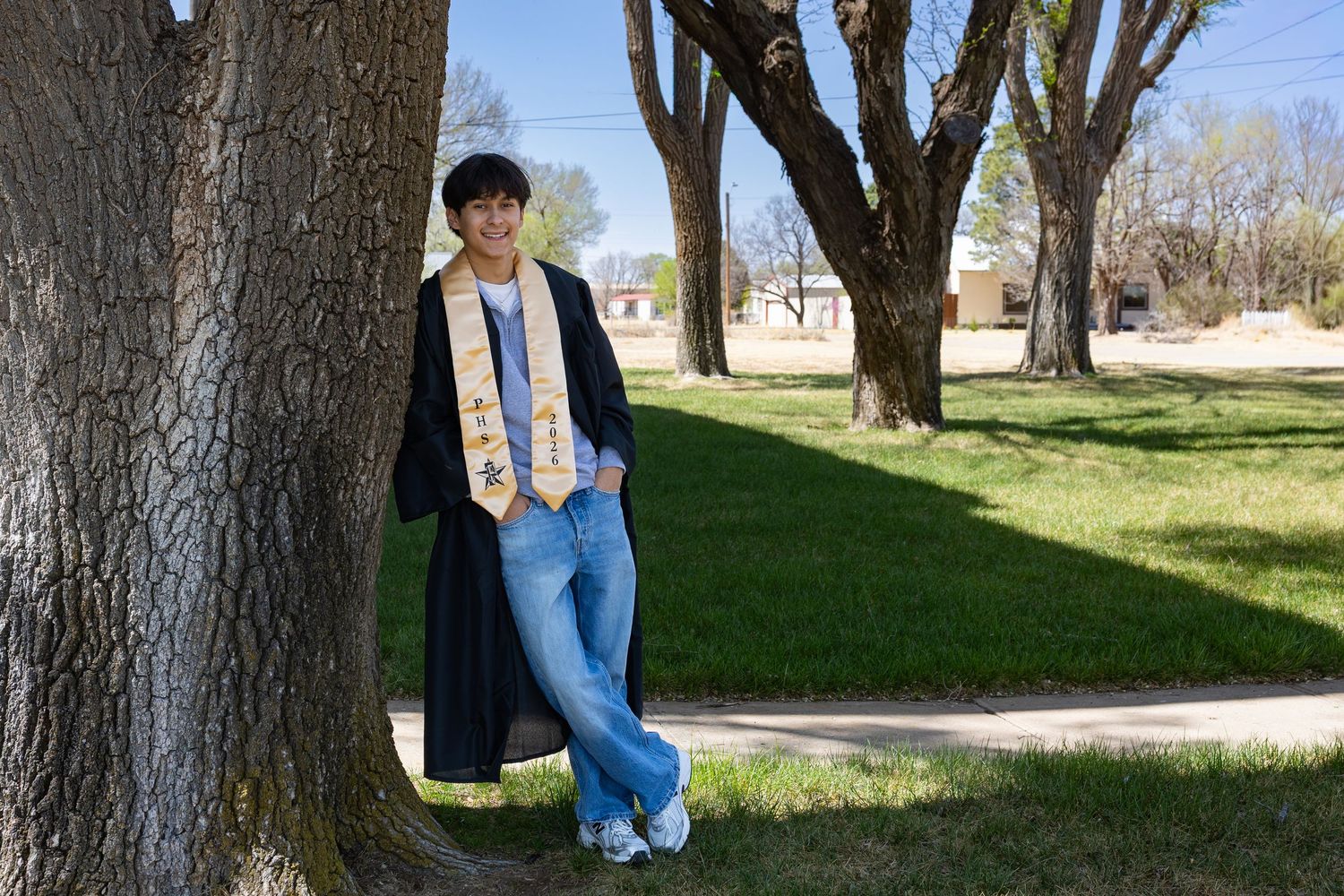 Graduate in black gown and gold stole leaning against a tree in a park for a senior portrait photo.