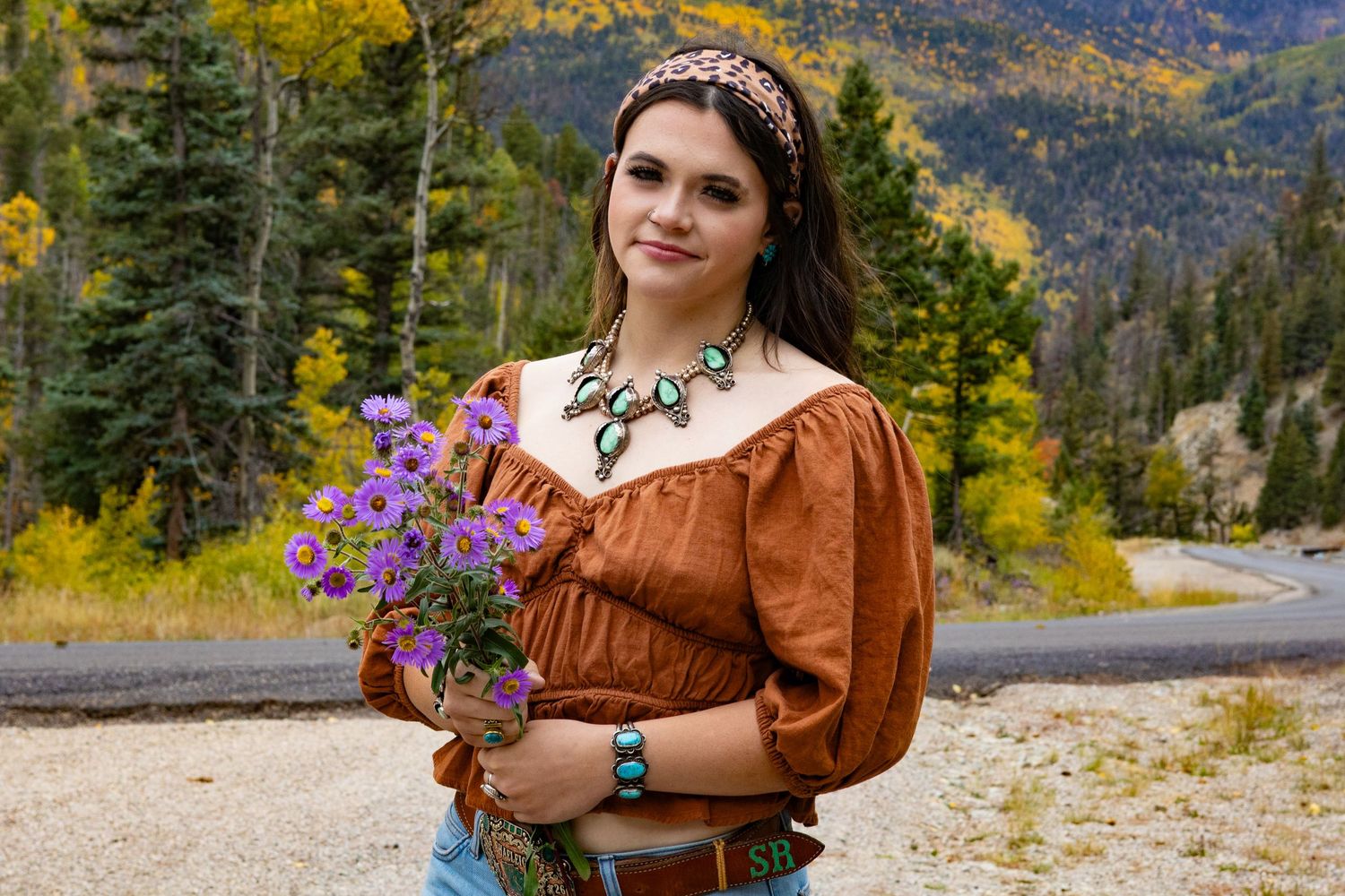 Young woman in rustic brown blouse holding purple wildflowers with turquoise jewelry amid autumn mountain scenery.