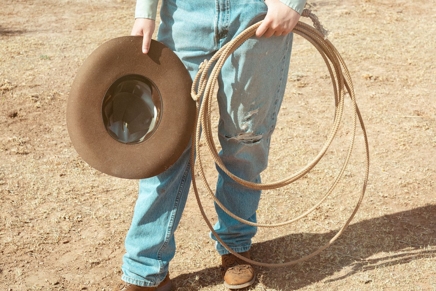 Cowboy holding a tan hat and lasso rope in sandy terrain.