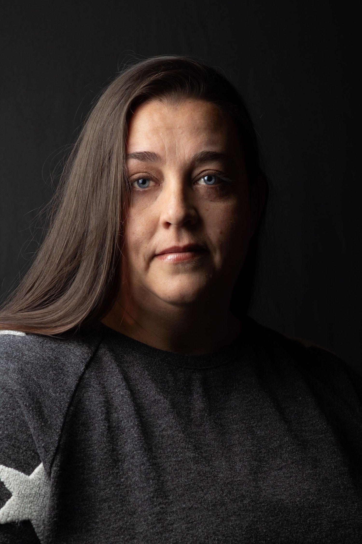 Woman with long brown hair and blue eyes in serious dark studio portrait wearing gray star sweater.