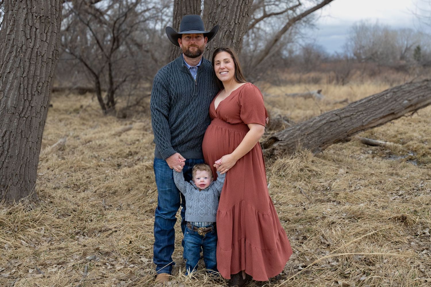 Family maternity portrait with cowboy hat dad, pregnant mom in rust dress, and toddler outdoors.