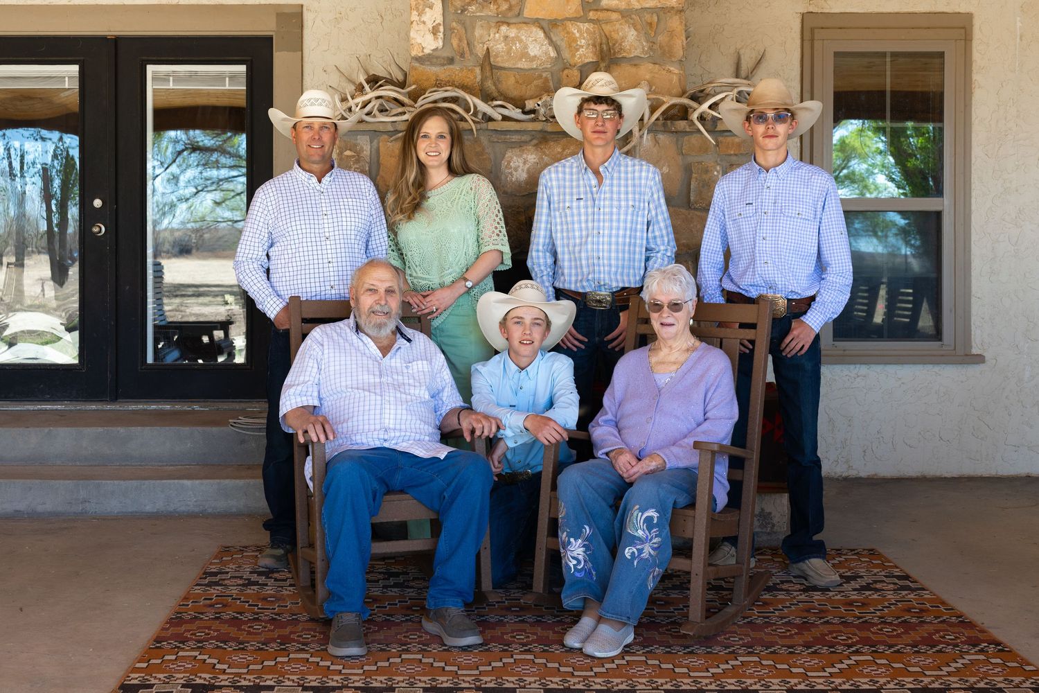 Seven family members pose on a rustic ranch porch, with four wearing cowboy hats in western wear.