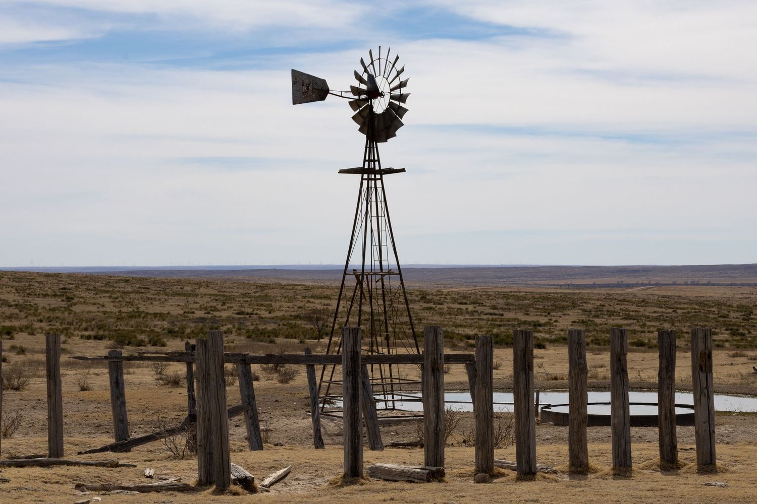 Vintage windmill and wooden fence posts on open prairie under cloudy sky.