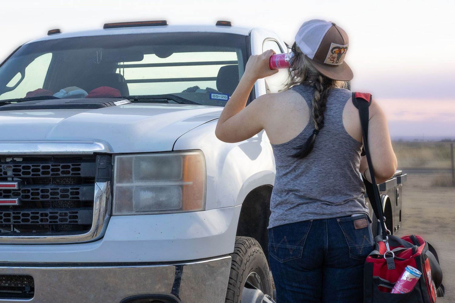 Woman in gray tank top and cap leaning against a silver GMC truck at dusk in an open field.