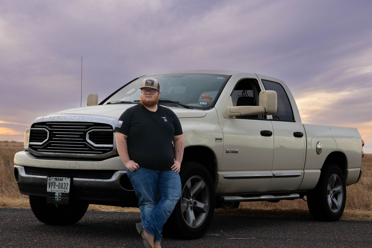 Man leaning against white Dodge Ram truck on open road beneath colorful purple and orange sunset sky.