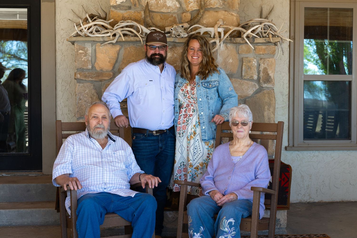 Four family members pose on a stone ranch porch, two seated in wooden chairs and two standing behind.