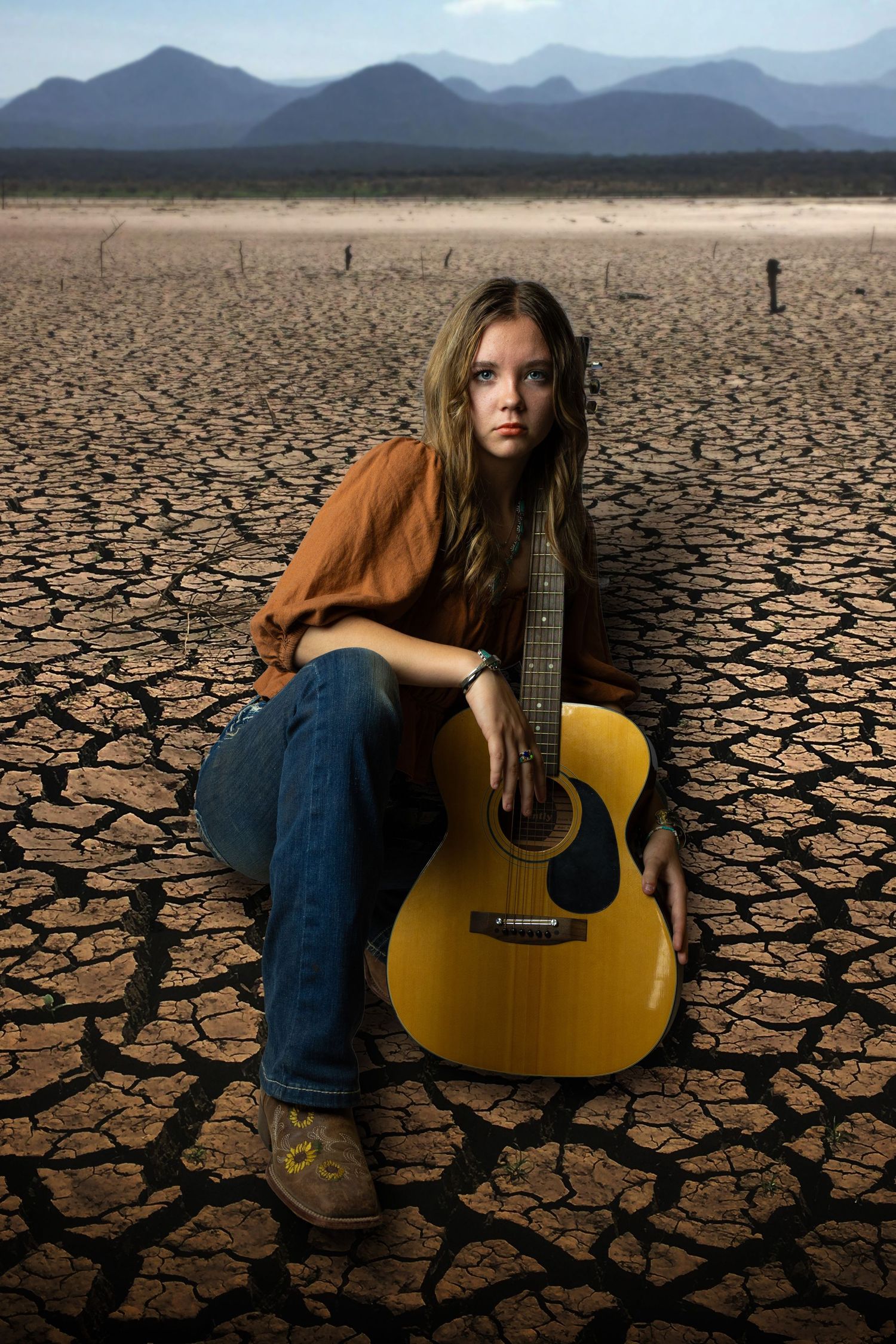 Young woman with long hair sits on cracked desert earth holding a yellow acoustic guitar with mountains behind.
