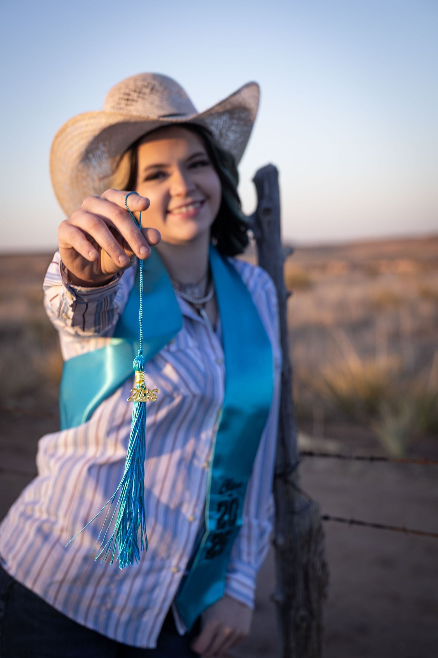 Smiling young woman in cowboy hat holding graduation tassel toward camera at golden hour near barbed wire fence.