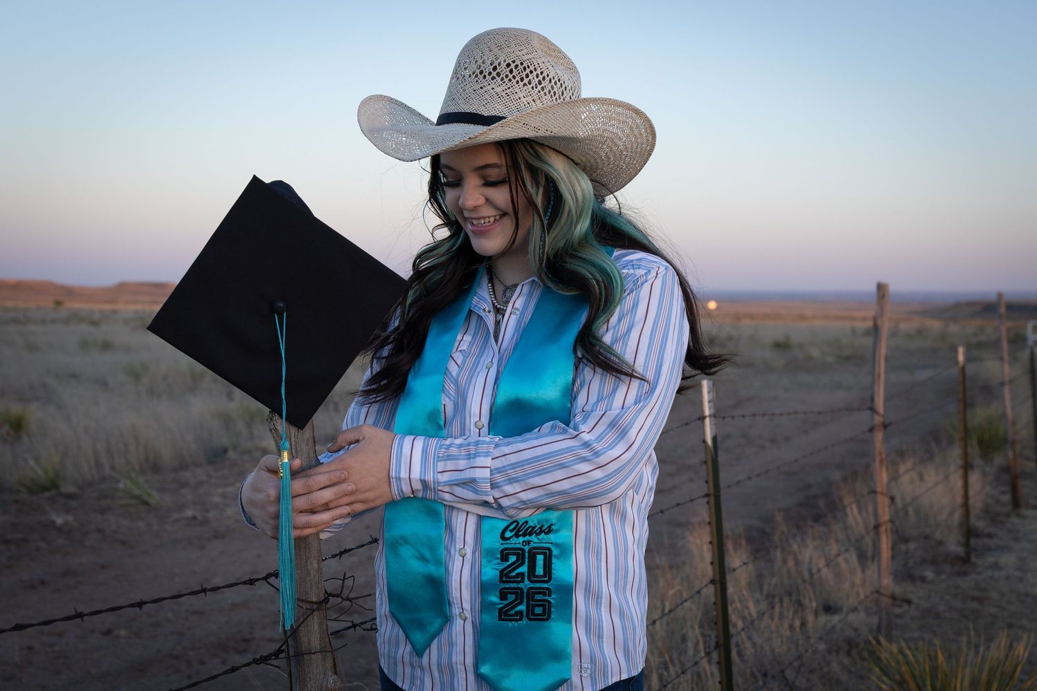 Young woman in cowboy hat examines Class of 2025 graduation stole at dusk near rustic barbed wire fence.
