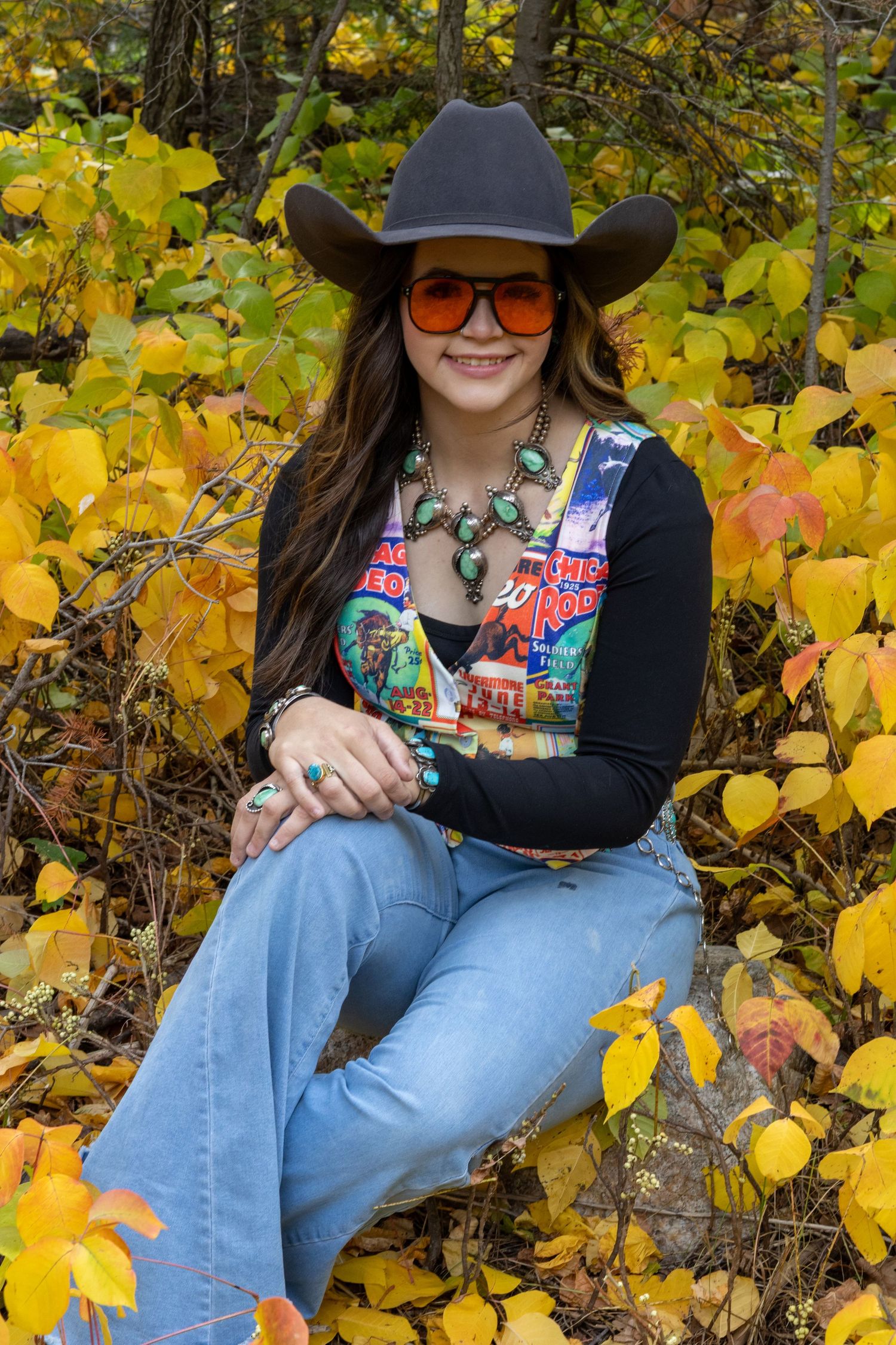 Smiling woman in cowboy hat and orange sunglasses sitting among vibrant yellow autumn foliage wearing turquoise jewelry.