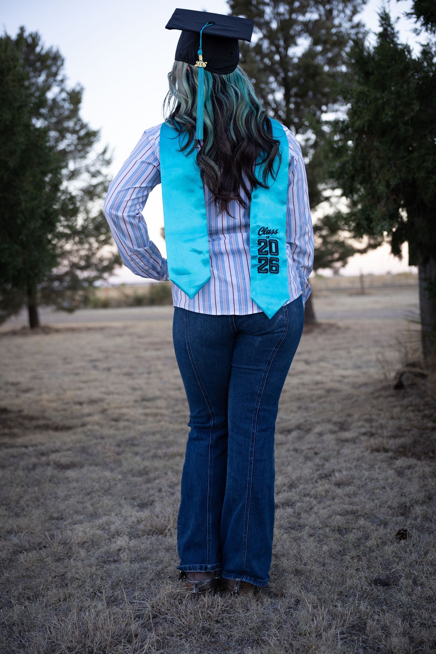 Back view of graduate with teal hair highlights wearing Class of 2025 stole and graduation cap outdoors.