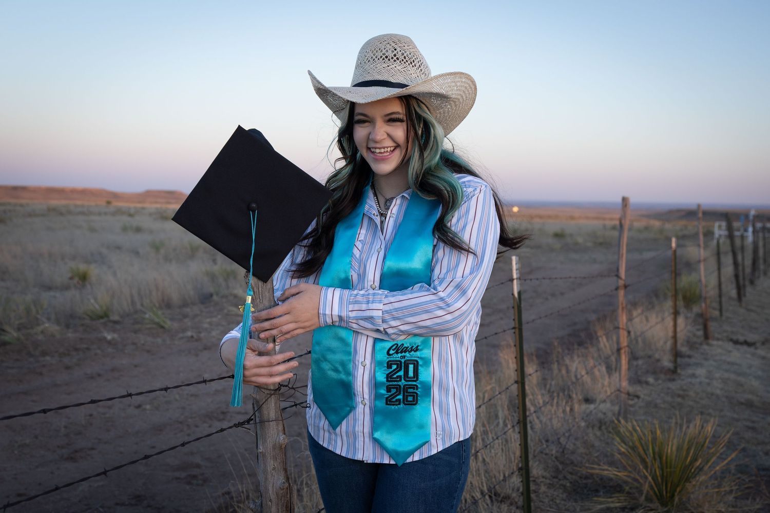 Smiling graduate in cowboy hat and turquoise Class of 2025 stole holds cap in open prairie at dusk.