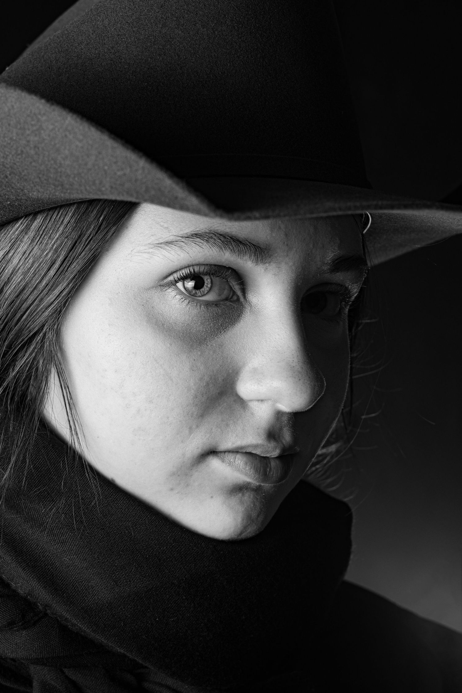 Dramatic black and white close-up portrait of young woman wearing wide-brim cowboy hat with intense gaze.